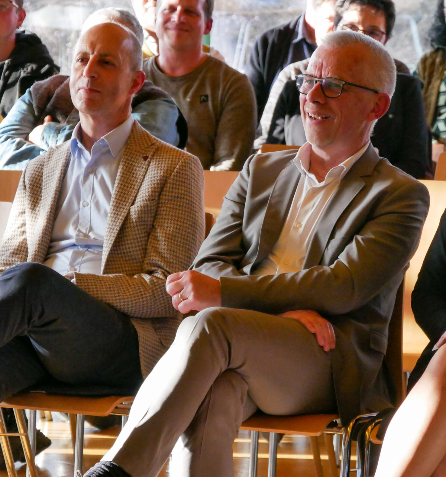 Zwei Männer in Anzügen, Alois Kohl und Johannes Blaurock, sitzen auf Holzstühlen bei einer Veranstaltung der Stiftung Haus Lindenhof. Sie sind von anderen in Freizeitkleidung umgeben, lächelnd und engagiert, während Sonnenlicht durch die Fenster hinter ihnen strömt.