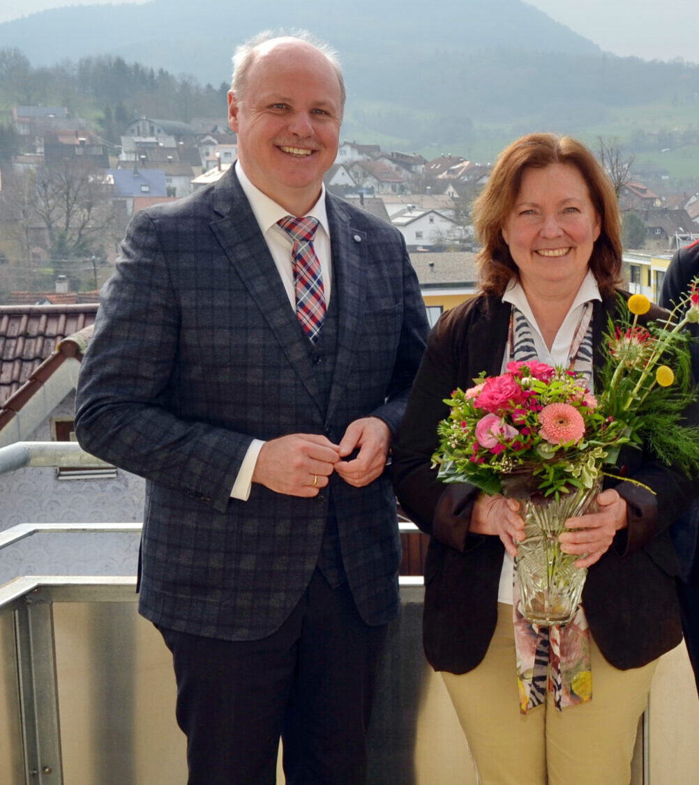 Magdalene Rupp im karierten Anzug und eine Frau mit einem Strauß bunter Blumen lächeln auf dem Balkon. Der malerische Blick auf die Stadt und die sanften Hügel bieten eine perfekte Kulisse, während Magdalene ihren Ruhestand mit Wärme und Freude feiert.