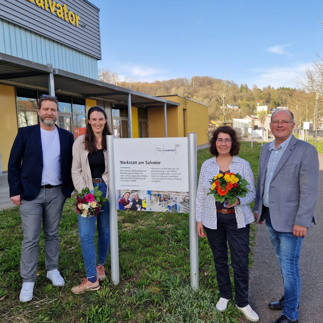 Vier Menschen stehen lächelnd im Freien neben einem Schild mit der Aufschrift „Werkstatt am Salvator“ und feiern einen Leitungswechsel. Zwei halten Blumensträuße in den Händen, um den Anlass zu feiern. Das Gebäude hinter ihnen trägt ebenfalls das Wort „Salvator“, und eine Rasenfläche und Bäume runden die ruhige Kulisse ab.