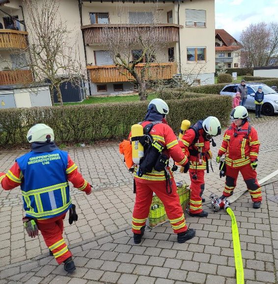 Vier Feuerwehrmänner in roten Uniformen und Helmen bereiten auf einem gepflasterten Platz in der Nähe eines Pflegeheims in Niederstotzingen die Löschausrüstung vor. Ein Feuerwehrmann trägt eine blaue Weste, während sie sich für die Feuerwehrübung vorbereiten. Sie stehen neben einem aufgerollten gelben Schlauch und stellen sicher, dass alles einsatzbereit ist.