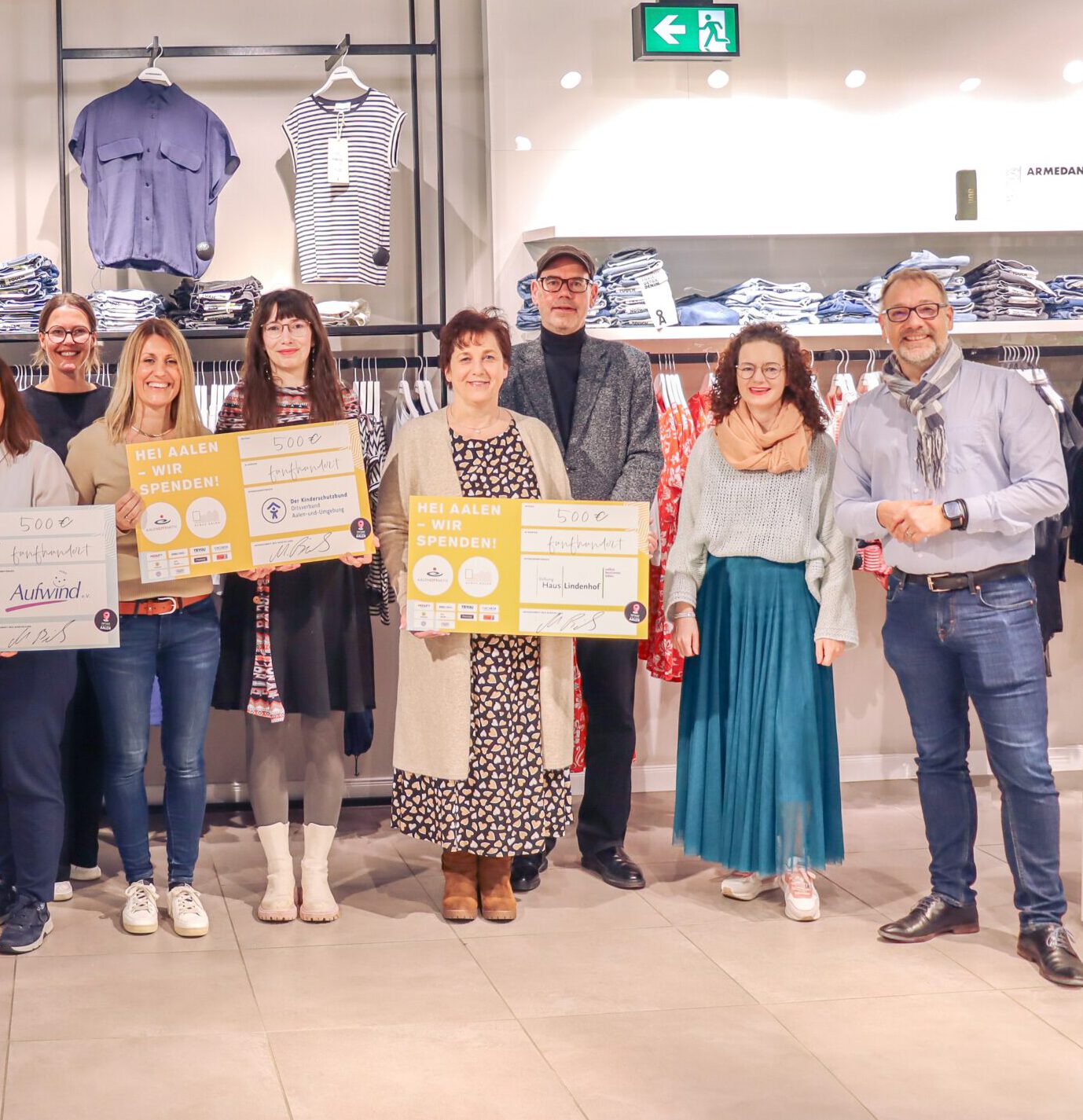 Eine Gruppe von Menschen steht in einem Bekleidungsgeschäft in Aalen für eine Spendenübergabe und hält große Schecks über je 500 Euro in den Händen. Sie lächeln in die Kamera, während auf Ständern hinter ihnen KUBUS-Kleidung ausgestellt ist.