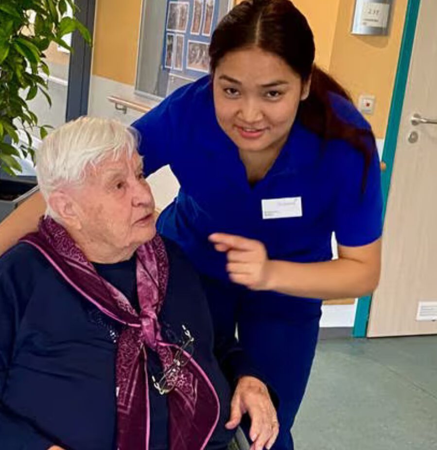 Eine Pflegekraft im blauen OP-Kittel der Stauferklinik lächelt, während sie einer älteren Frau mit weißem Haar hilft, die einen dunklen Pullover und einen kastanienbraunen Schal trägt. Sie sind drinnen, mit einer Pflanze und einer Tür im Hintergrund, und teilen einen Moment der Wärme und Fürsorge.
