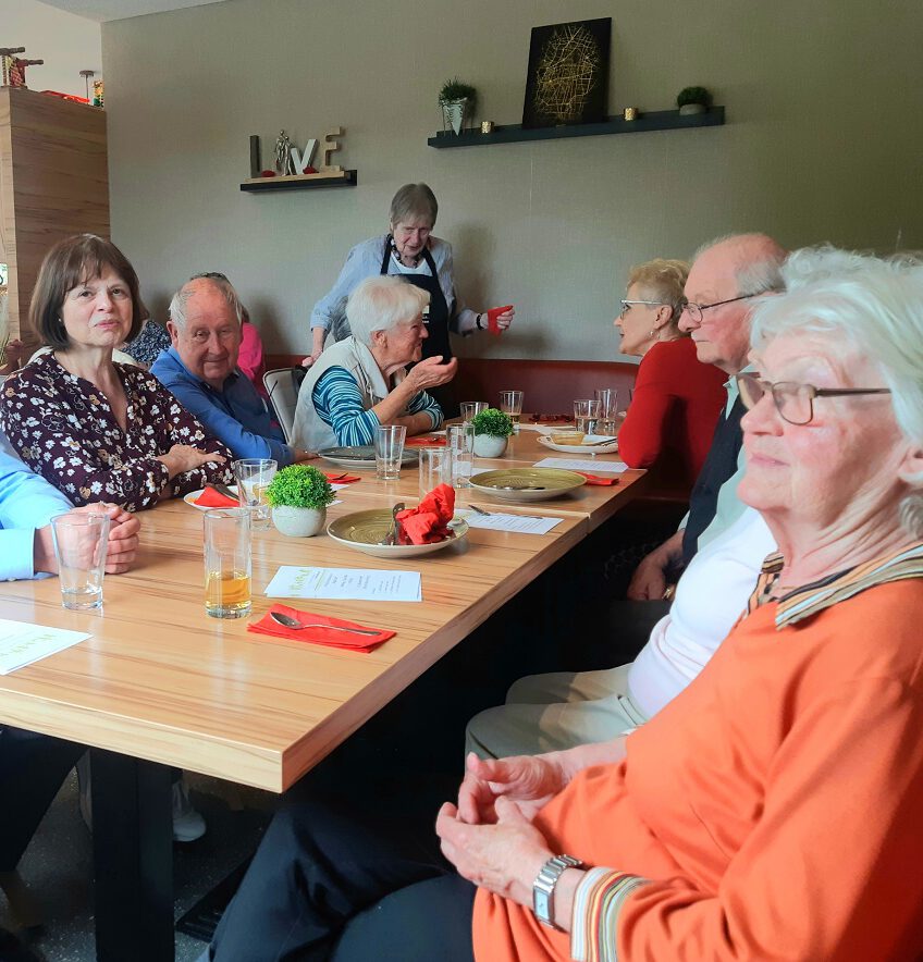 Eine Gruppe älterer Menschen sitzt um einen Tisch im Esszimmer und genießt einen Gelungener Beuremer Mittagstisch. Sie unterhalten sich angeregt, umgeben von kleinen Pflanzen und Getränken, an der Wand hängt ein dekoriertes Regal. Die Atmosphäre ist warm und gesellig.