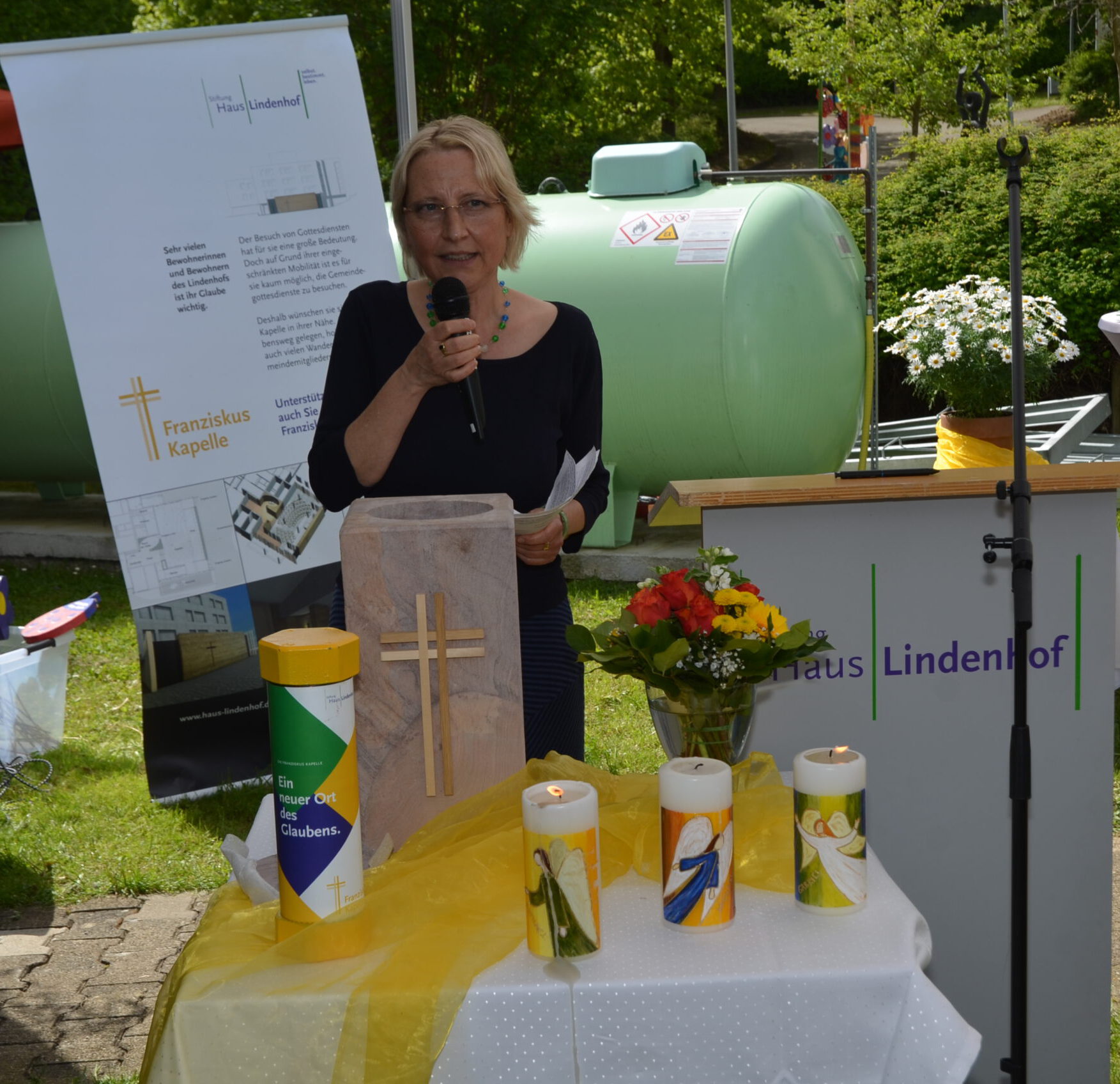 Eine Frau steht mit einem Mikrofon in der Hand an einem Podium im Freien. Vor ihr steht ein Tisch mit dekorierten Kerzen, einer Blumenvase und einer Holzkiste mit einem Kreuz, das die Grundsteinlegung der Franziskus-Kapelle markiert. Hinter ihr sind Plakate und ein grüner Tank zu sehen.