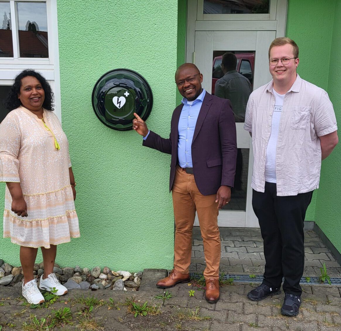 Drei Menschen stehen vor dem Bischof-Sproll-Haus, einem leuchtend grünen Gebäude. Die Person in der Mitte zeigt auf einen montierten Heubach-Defibrillator. Alle lächeln, eine trägt Kleid und Turnschuhe, eine andere Blazer und Hose und die dritte Hemd und Jeans.