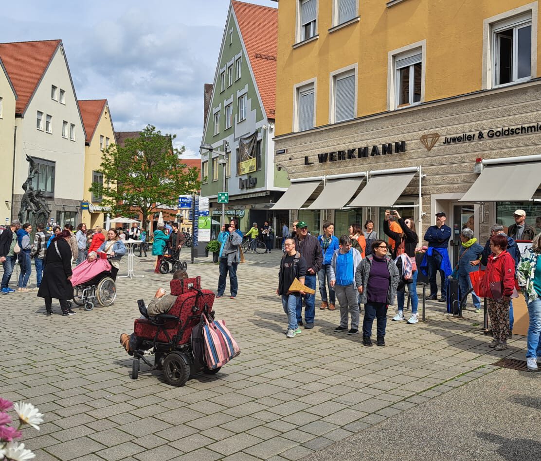 In Ellwangen spielt sich ein geschäftiges Straßenbild ab, wo Menschen auf einem lebhaften Platz gehen und stehen. Im Vordergrund stehen zwei Rollstuhlfahrer, die den Gleichstellungstag symbolisieren, während sich um sie herum verschiedene Fußgänger verteilen. Farbenfrohe Gebäude, darunter ein Juweliergeschäft, säumen den Hintergrund unter einem teilweise bewölkten Himmel.