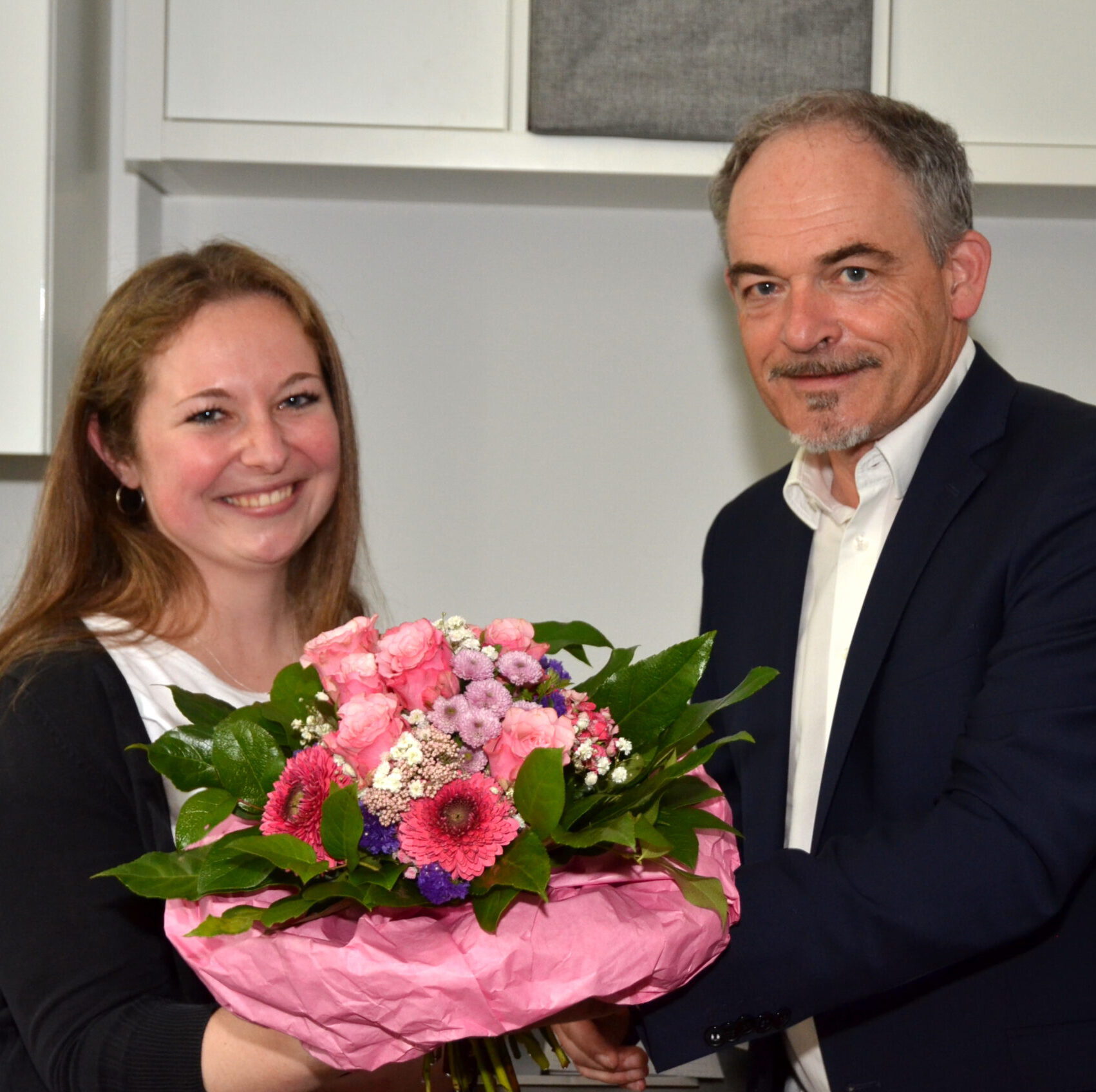 Eine Frau lächelt und hält einen bunten Blumenstrauß in rosa Papier eingewickelt in der Hand, um einen Leitungswechsel zu feiern. Neben ihr lächelt ein Mann in dunkler Jacke leicht. Sie stehen in einem gut beleuchteten Raum mit weißen Schränken im Hintergrund.