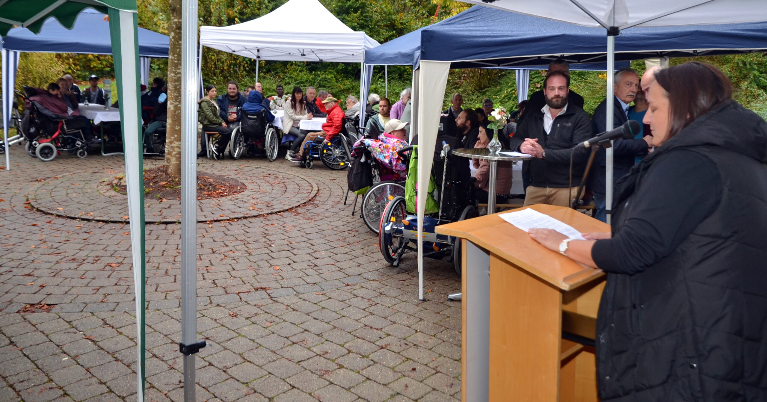 Eine Frau spricht auf einem Podium unter einem Baldachin im Bischof Ketteler Haus. Menschen sitzen an Tischen und in Rollstühlen, geschützt durch Zelte auf dem Kopfsteinpflasterweg, und feiern das 25-jährige Jubiläum. Der Bereich ist von Grün umgeben und schafft eine integrative Versammlung im Freien.