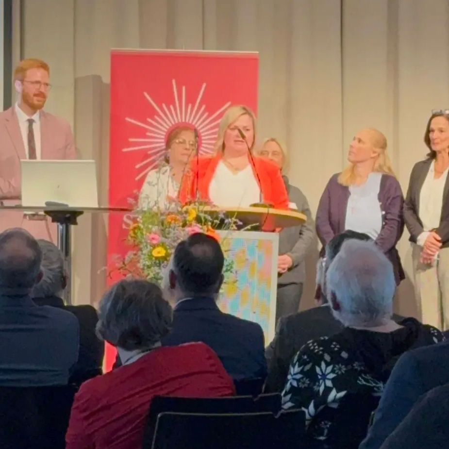 Eine Gruppe von Menschen steht hinter einem Podium, auf dem eine Frau in einem roten Blazer spricht. Ein Blumenarrangement schmückt das Podium, während sie an „10 Jahre Sozialstation St. Martin“ erinnert. Im Vordergrund sitzt ein Publikum, im Hintergrund ist ein Banner mit einem Sonnenstrahlen-Design zu sehen.