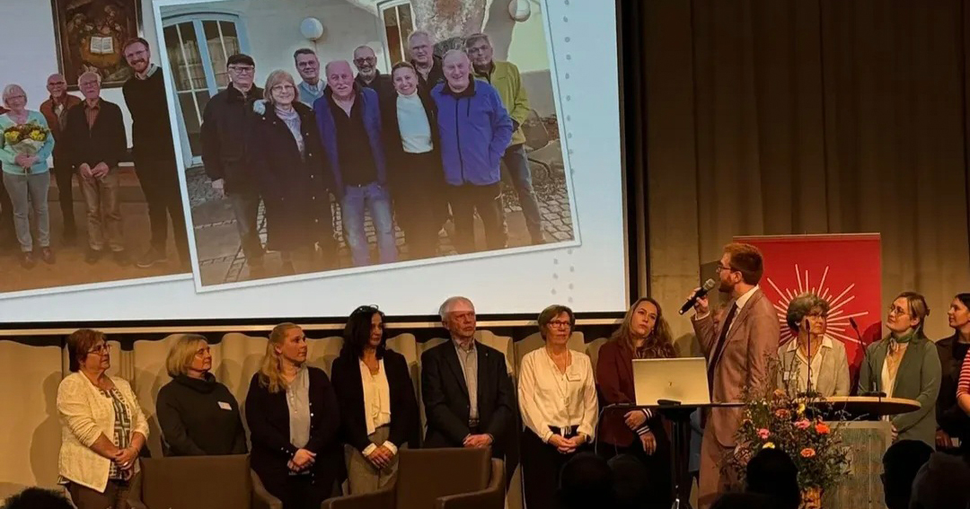 Eine Gruppe von Menschen steht auf einer Bühne, eine Person spricht in ein Mikrofon. Hinter ihnen sind auf einer großen Leinwand Fotos einer anderen Gruppe zu sehen, die draußen posiert und das 10-jährige Jubiläum der Sozialstation St. Martin feiert. Ein Blumenarrangement schmückt einen Tisch neben einem roten Banner.