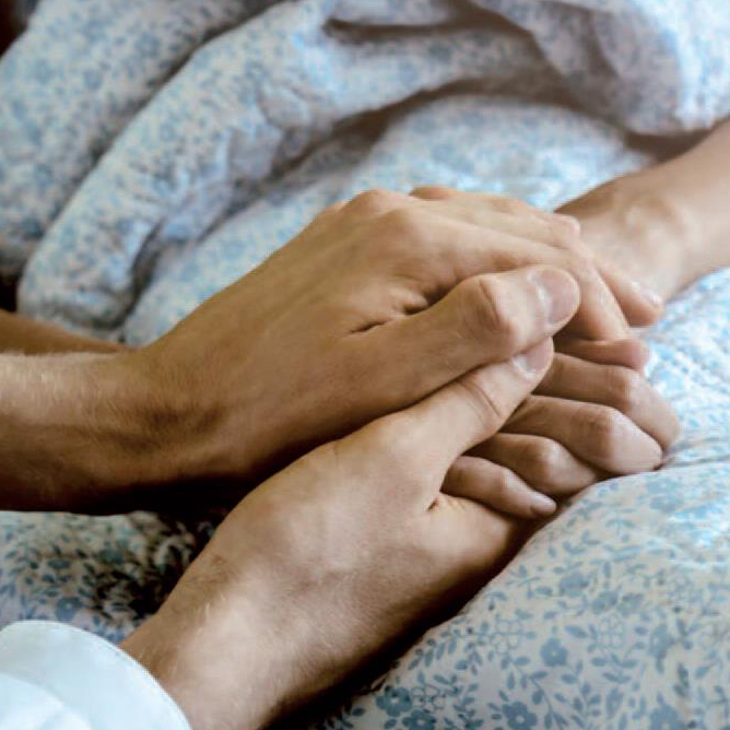 Zwei Hände halten sich sanft über einer blau-weißen, geblümten Decke und erinnern so an die Empathie, die im Hospiz Heidenheim herrscht. Die Nahaufnahme der gefalteten Hände vermittelt Trost, Unterstützung und Kameradschaft.