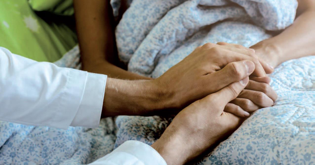 Eine Person mit einem weißen Ärmel hält die Hand einer anderen Person, die im Bett liegt, zugedeckt mit einer blau-weiß gemusterten Decke, und bietet Trost und Unterstützung, was an Zusammenkünfte wie den Fachtag Hospiz in Heidenheim erinnert.
