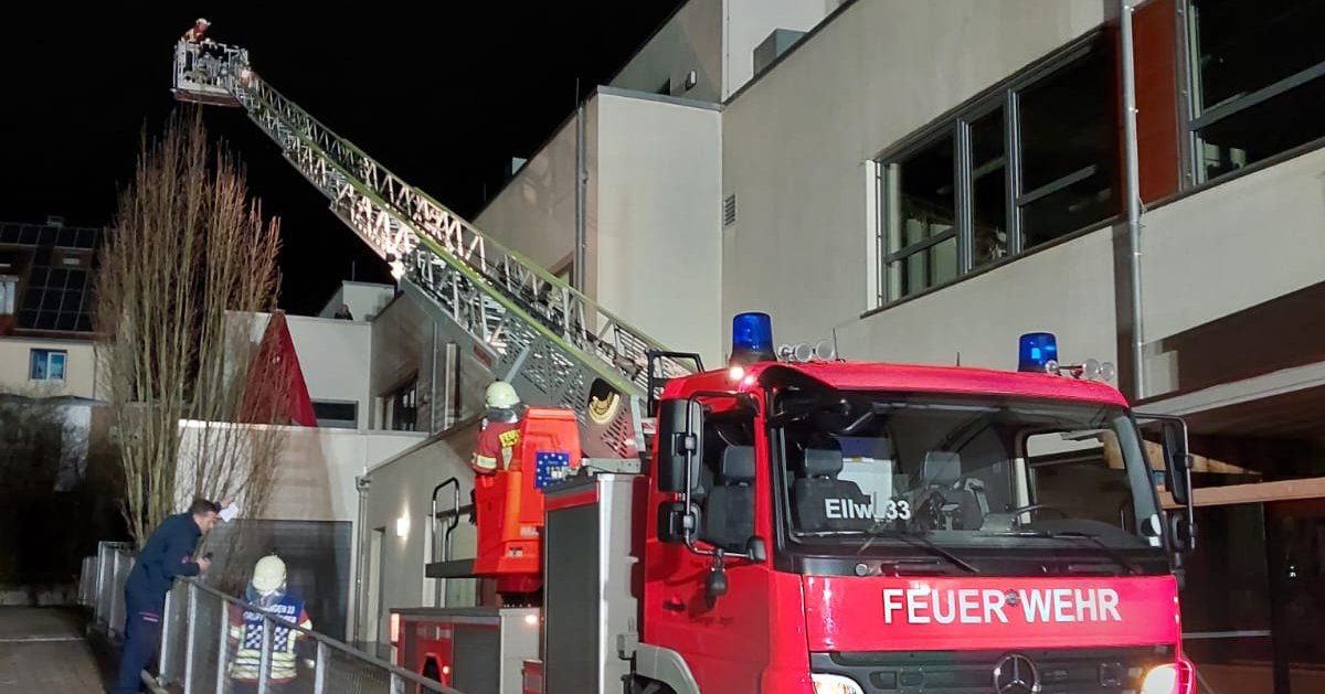 Ein Feuerwehrauto mit der Aufschrift „Feuerwehr“ fährt während einer nächtlichen Feuerwehrübung in Ellwangen seine Leiter aus. Feuerwehrleute sind aktiv im Einsatz: Einer steigt die Leiter hinauf, während andere am Boden bleiben und die Situation aufmerksam überwachen.