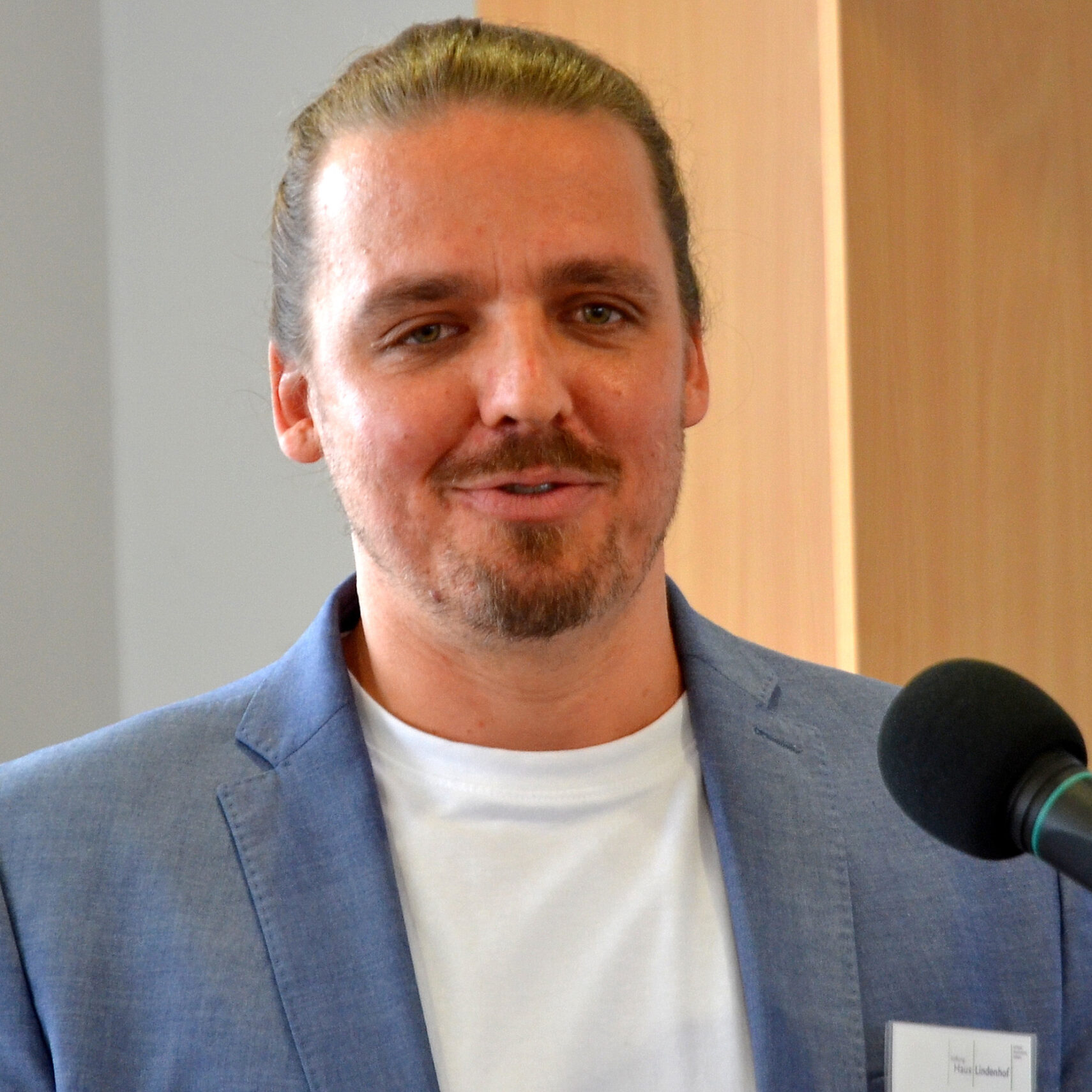 Benedikt Saur, der neue Leiter der Christophorus Werkstatt, trägt einen Bart und Schnurrbart und spricht im hellblauen Blazer und weißen Hemd ins Mikrofon. Mit zurückgebundenem Haar steht er in Innenräumen nahe einer Holzvertäfelung.