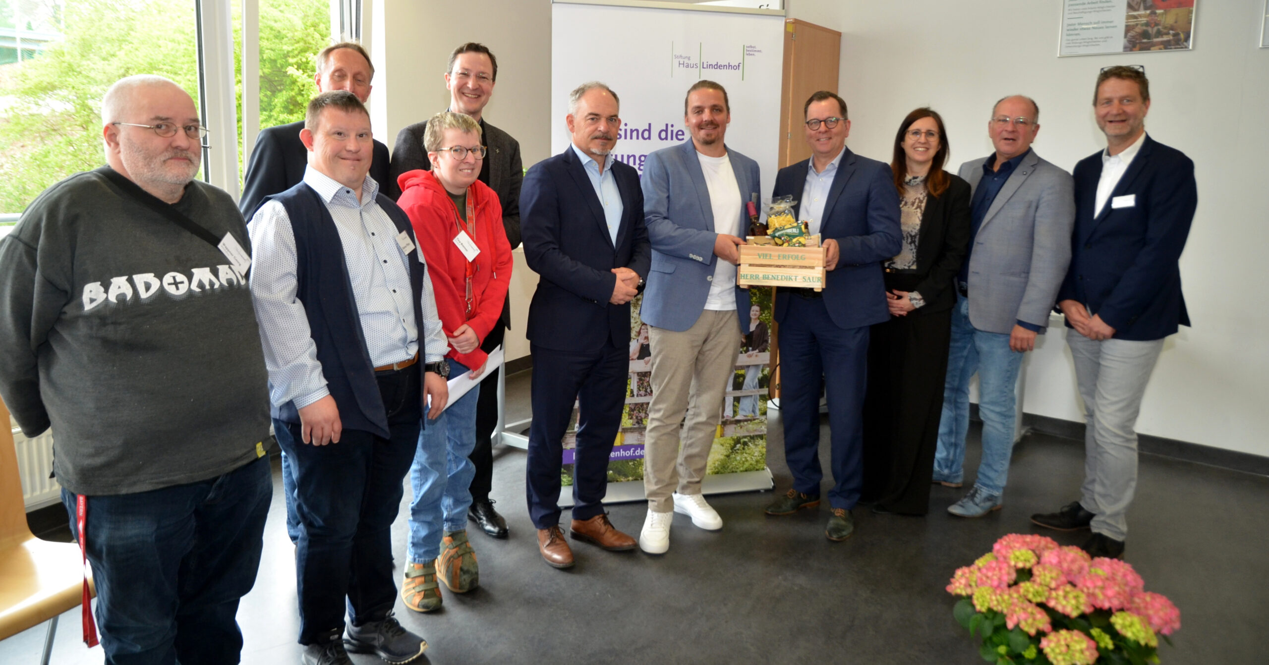 Eine Gruppe von elf Personen steht im Innenbereich und lächelt in die Kamera. In der Mitte hält Benedikt Saur, der neue Leiter der Christophorus Werkstatt, einen Geschenkkorb und eine Plakette. Im Hintergrund sind ein Banner und ein Fenster mit Grünpflanzen zu sehen.
