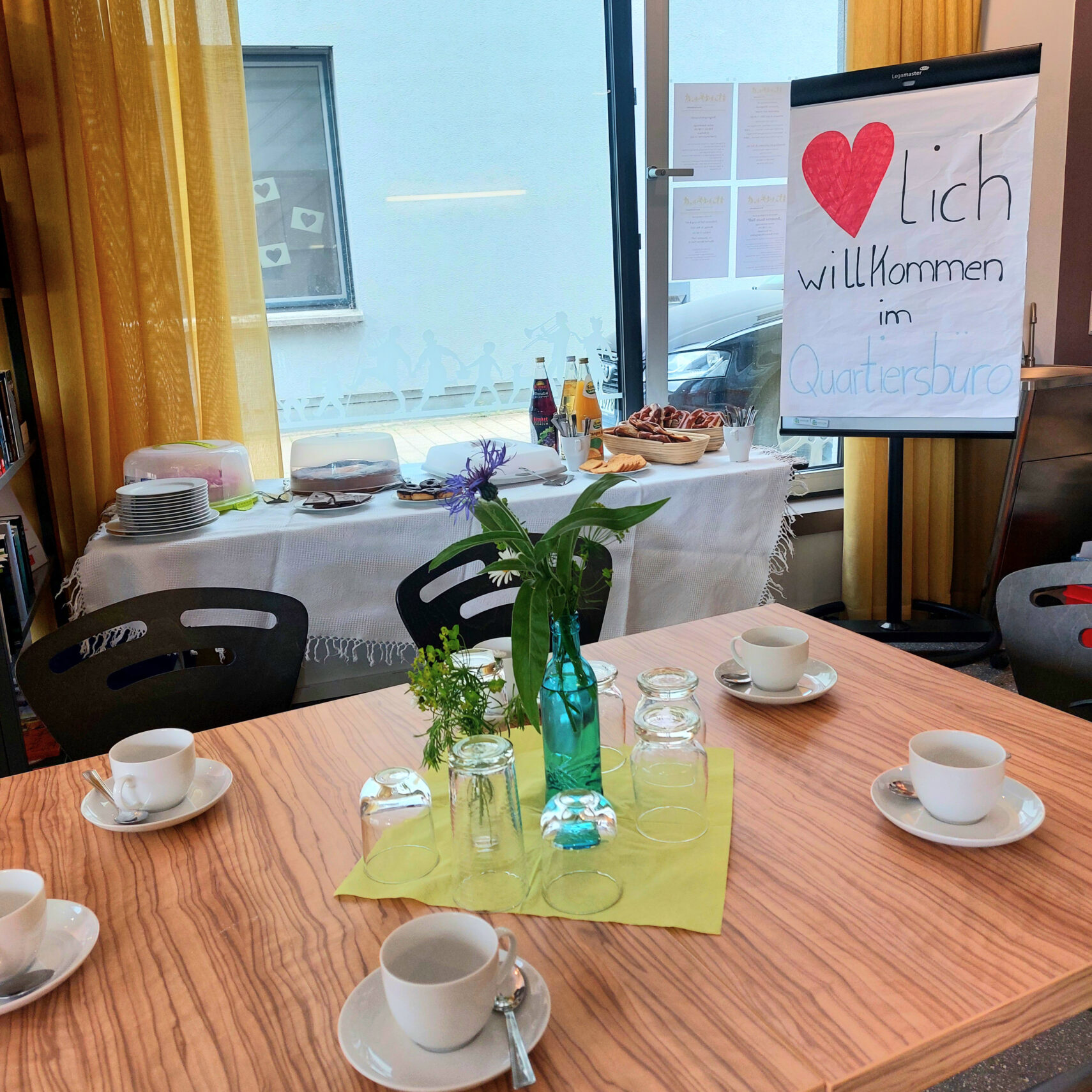 Ein gedeckter Tisch mit Tassen, Untertassen, Tellern und einer Blumenvase. Im Hintergrund begrüßt ein Buffet mit Getränken und Gebäck die Gäste im Café Kunterbunt; auf einem Schild mit einem roten Herzen steht „Herzlich Willkommen im Quartiersbüro“.