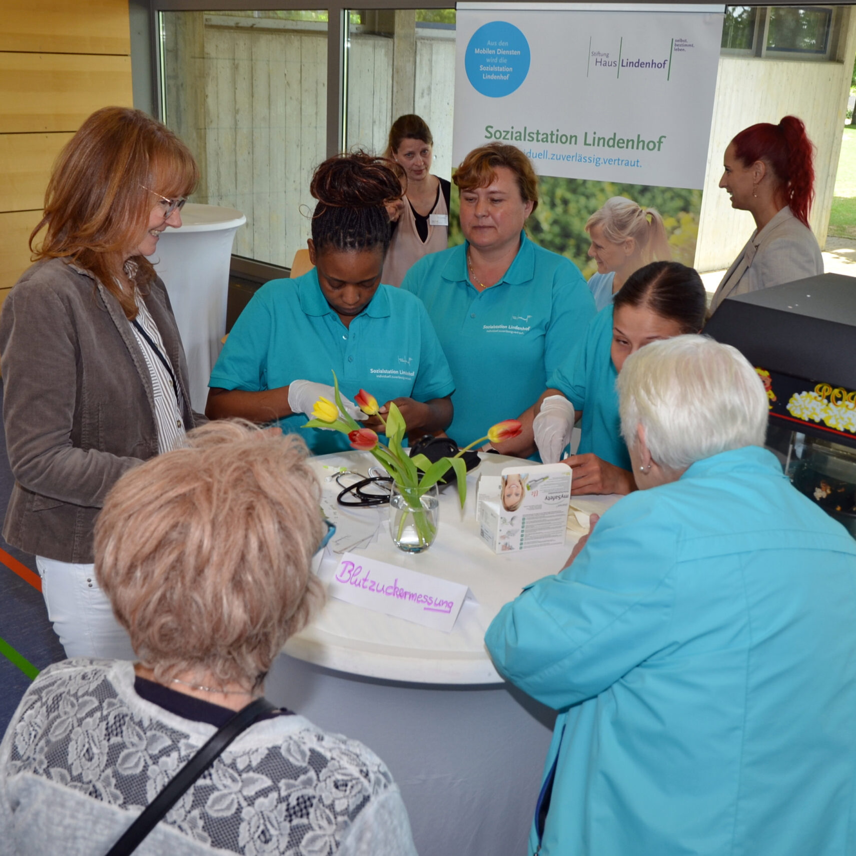 Eine Gruppe von Frauen, einige in türkisfarbenen Uniformen, steht auf der Pflegemesse in Waldstetten um einen runden Tisch mit Blumen, Broschüren und einem Schild. Sie scheinen bei dieser Indoor-Veranstaltung an einer Diskussion oder Demonstration teilzunehmen.