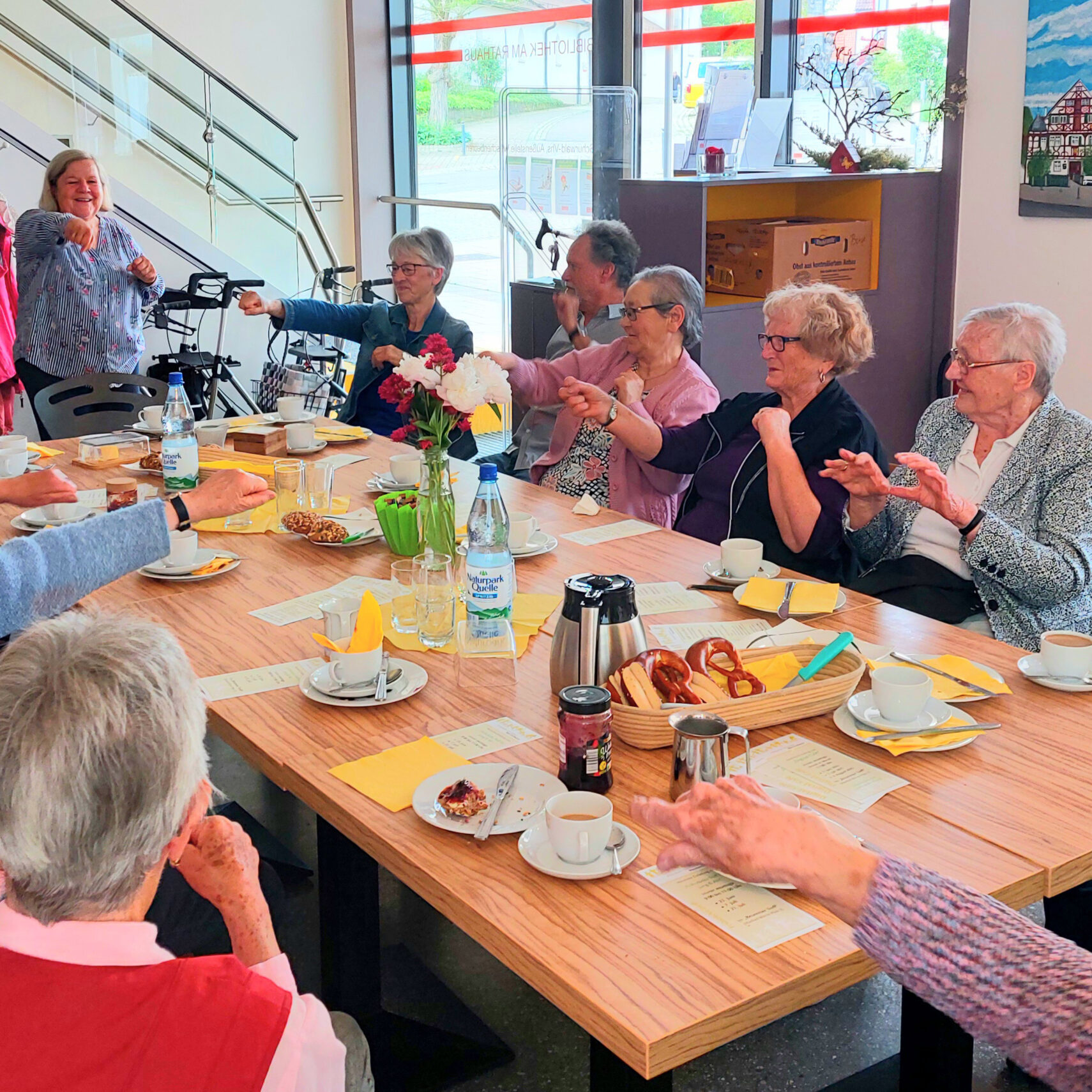 Beim Frühstückstreff in Wäschenbeuren sitzt eine Gruppe älterer Menschen bei Kaffee, Gebäck und Blumen um einen Tisch, lacht und zeigt fröhlich aufeinander in einem hellen, freundlichen Raum.