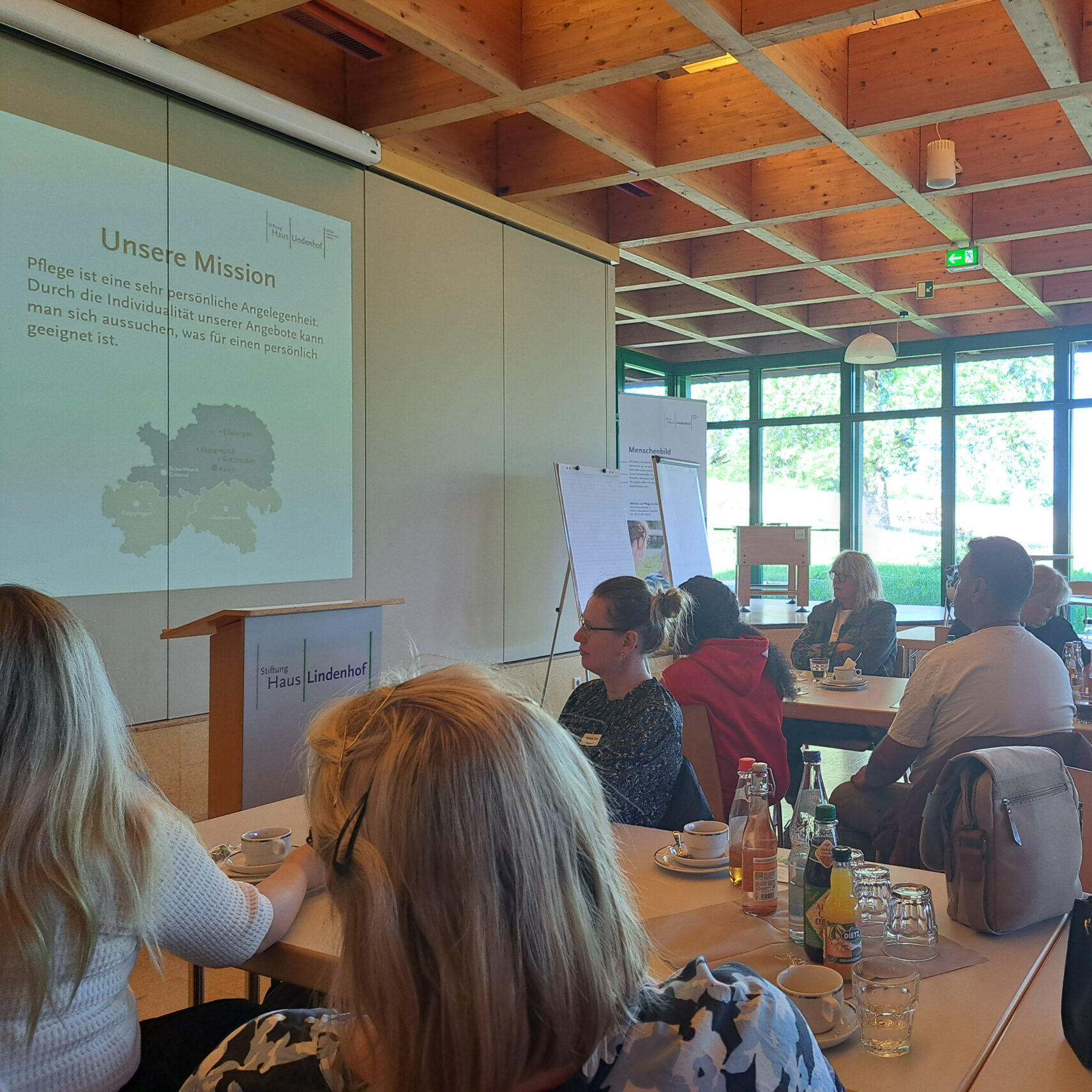Eine Gruppe von Menschen, darunter auch neue Mitarbeitende, sitzt an Tischen in einem hellen Konferenzraum für eine Einführungsveranstaltung und blickt aufmerksam auf einen Präsentationsbildschirm mit dem deutschen Text „Unsere Mission“. Getränke und Unterlagen stehen auf den Tischen.