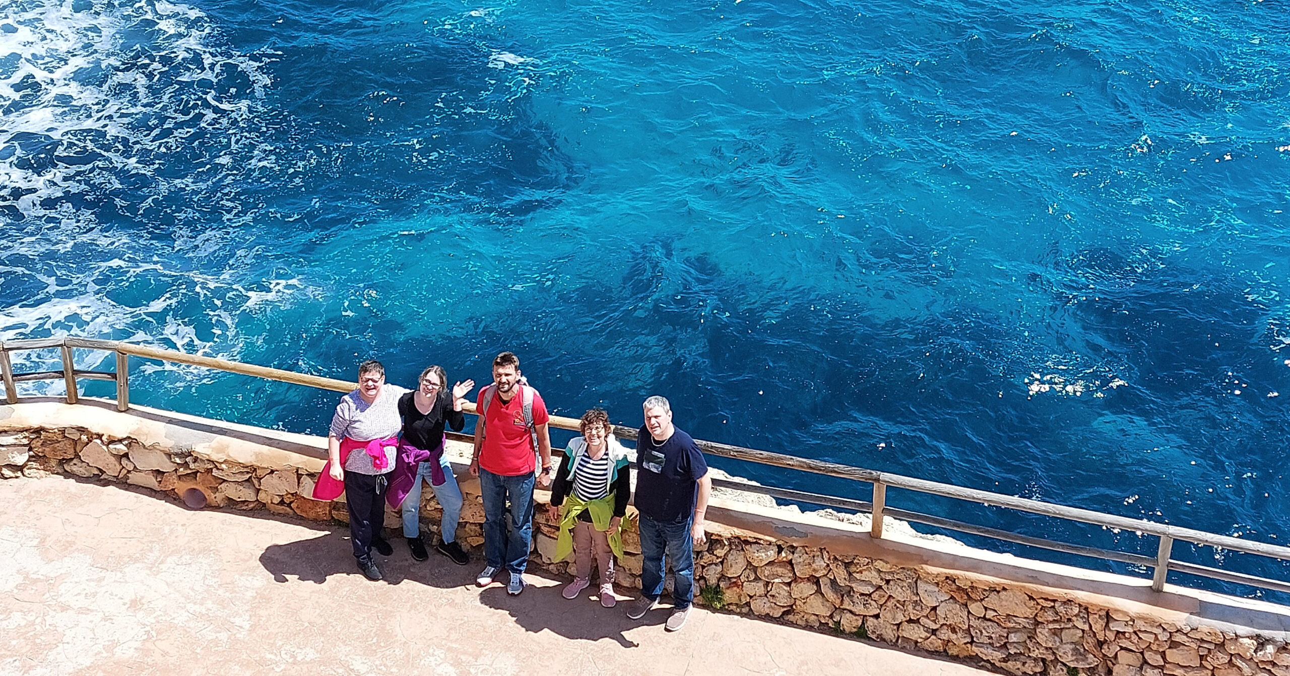 Sechs Menschen stehen auf einem Steinweg am blauen Meer auf Mallorca und lächeln in die Kamera. Ein Holzgeländer trennt sie vom glitzernden Wasser – ein Moment, der an ein Treffen mit Freunden in der WG Jakob-Uhlmann-Straße erinnert.