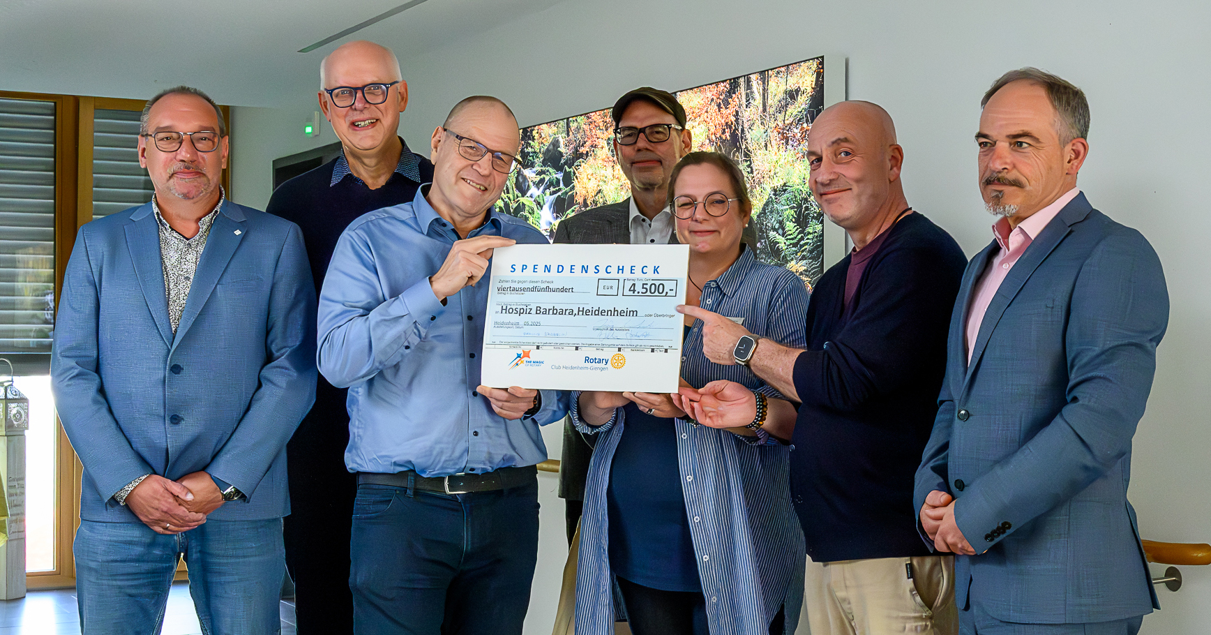 Sieben Personen stehen lächelnd im Innenbereich zusammen und halten einen Spendenscheck über 4.500 Euro für das Hospiz Barbara in Heidenheim in den Händen. Die Gruppe wirkt fröhlich und trägt elegante Freizeitkleidung.