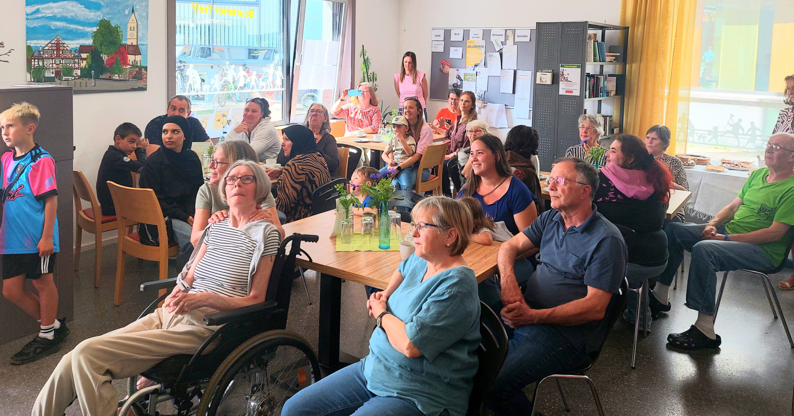 Eine bunt gemischte Gruppe von Menschen, darunter Kinder und Erwachsene, sitzt im hellen Gemeinschaftsraum des Café Kunterbunt mit seinen großen Fenstern, Anschlagtafeln und blumengeschmückten Tischen und schaut sich etwas außerhalb des Kamerabereichs an.