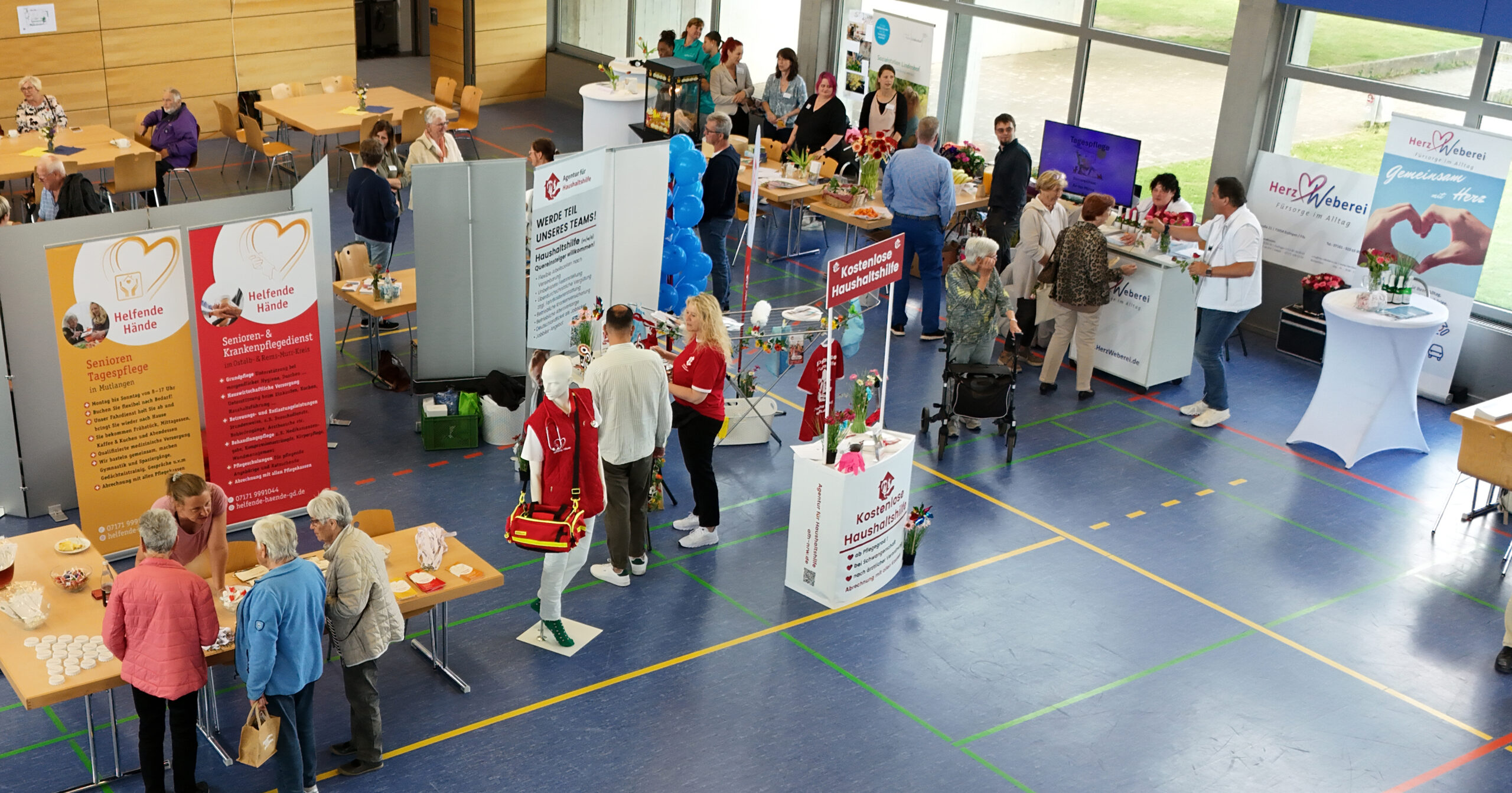 Eine Gesundheitsmesse in der Waldstettener Turnhalle. Besucher besuchen die Stände der Pflegemesse mit Bannern, Gesundheitsinformationen, medizinischer Ausrüstung und Luftballons. Einige Besucher sprechen mit Mitarbeitern in roten Jacken, andere erkunden Tische und Ausstellungen.