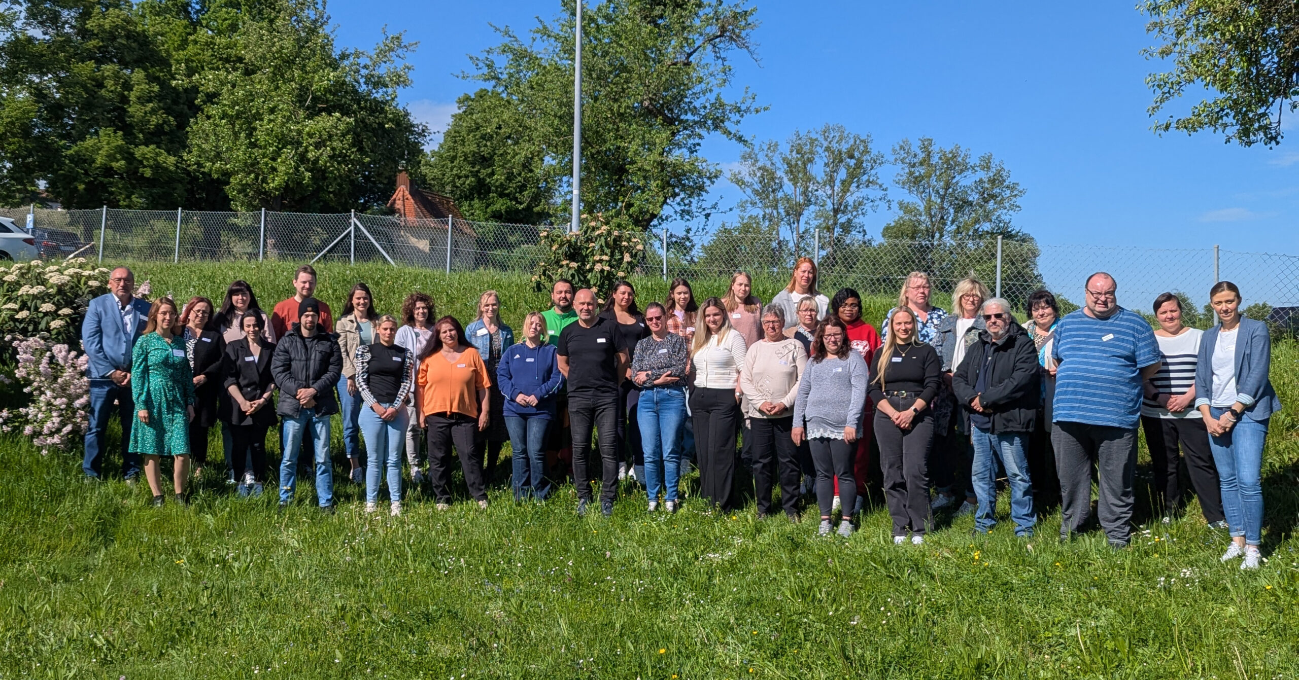 Eine große Gruppe von Menschen, darunter auch neue Mitarbeitende, steht während einer Einführungsveranstaltung im Freien auf einer Rasenfläche zusammen und posiert an einem sonnigen Tag mit Bäumen, Blumen und einem Zaun im Hintergrund für ein Foto.