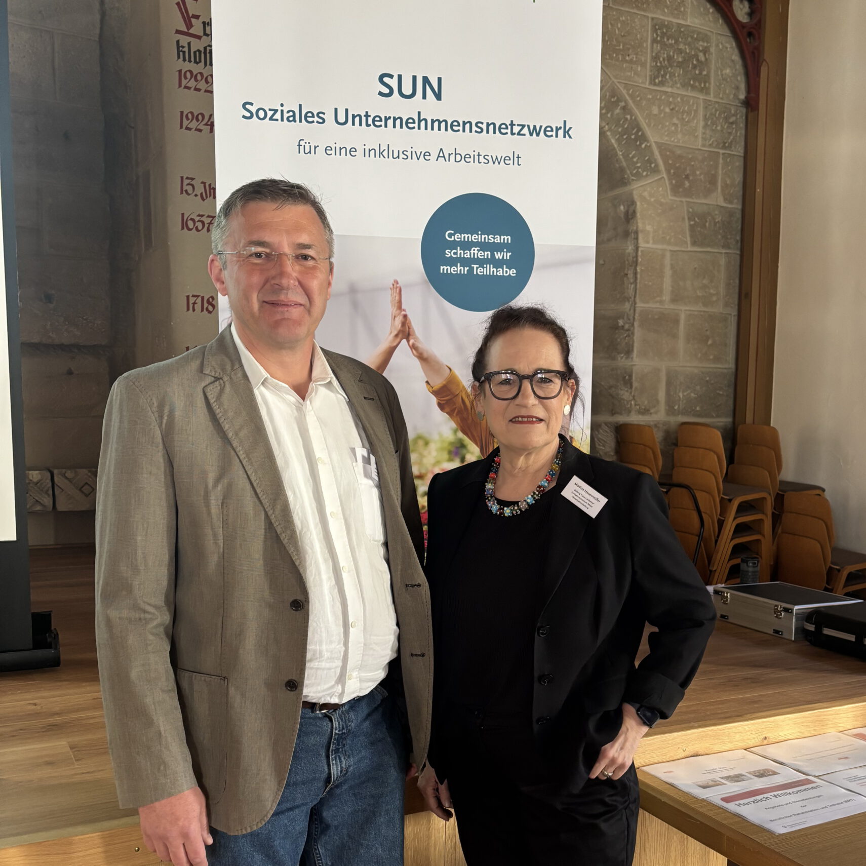 Ein Mann und eine Frau stehen drinnen vor einem Banner mit der Aufschrift SUN Soziales Unternehmensnetzwerk für eine inklusive Arbeitswelt, mit Stühlen, einer Steinmauer und einem automatisch gespeicherten Entwurf im Hintergrund.