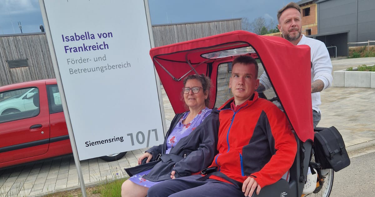 Eine Frau und ein Mann sitzen in einem roten Rikscha-Fahrrad, ein weiterer Mann hält den Lenker hinter ihnen. Sie stehen neben einem Schild mit der Aufschrift „Isabella von Frankreich“ vor einem modernen Gebäude, möglicherweise in der Nähe eines Förderbereichs.