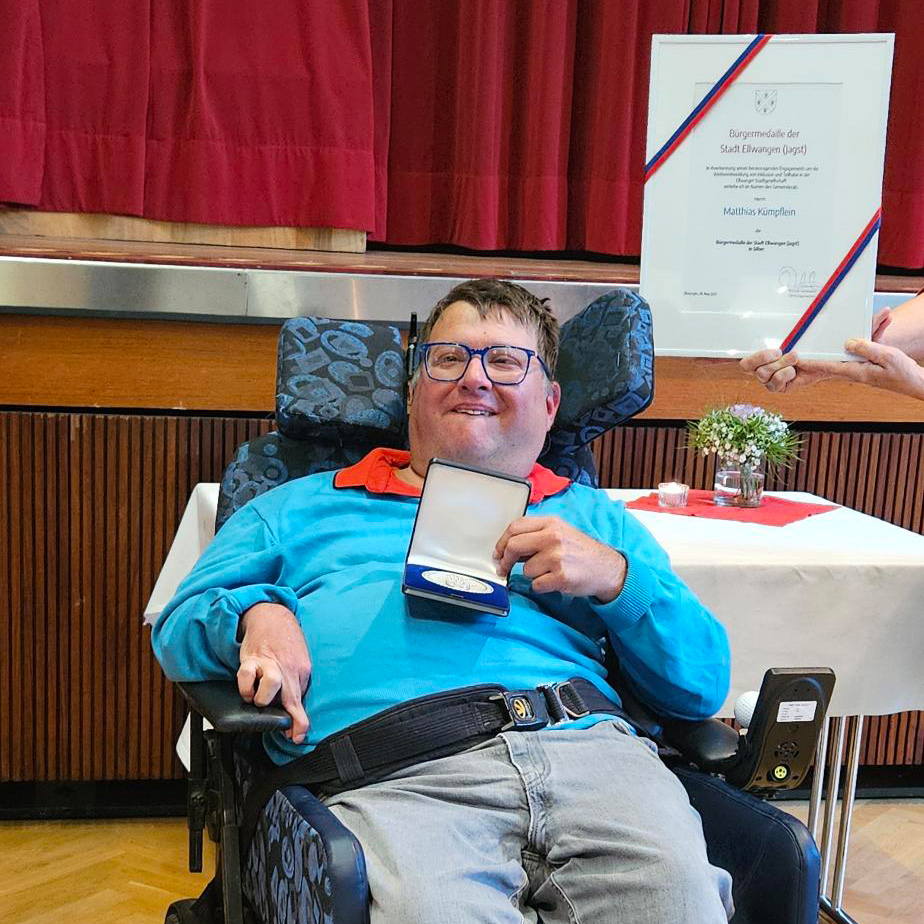 Ein lächelnder Matthias Kümpflein im Rollstuhl hält eine geöffnete Ellwanger Bürgermedaille in der Hand. Hinter ihm sind eine gerahmte Urkunde und eine Hand, die Blumen überreicht, zwischen roten Vorhängen und einer Holzwand zu sehen.