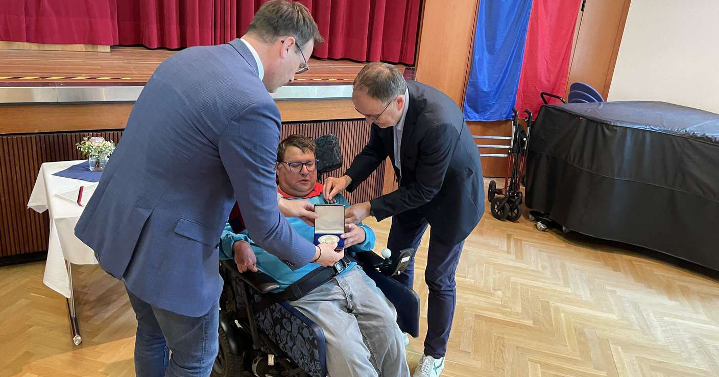 Zwei Männer in Anzügen überreichen dem im Rollstuhl sitzenden Matthias Kümpflein die Ellwanger Bürgermedaille während einer Indoor-Zeremonie mit roten Vorhängen und Holzfußboden. Der Empfänger zeigt sich gerührt von der Anerkennung.
