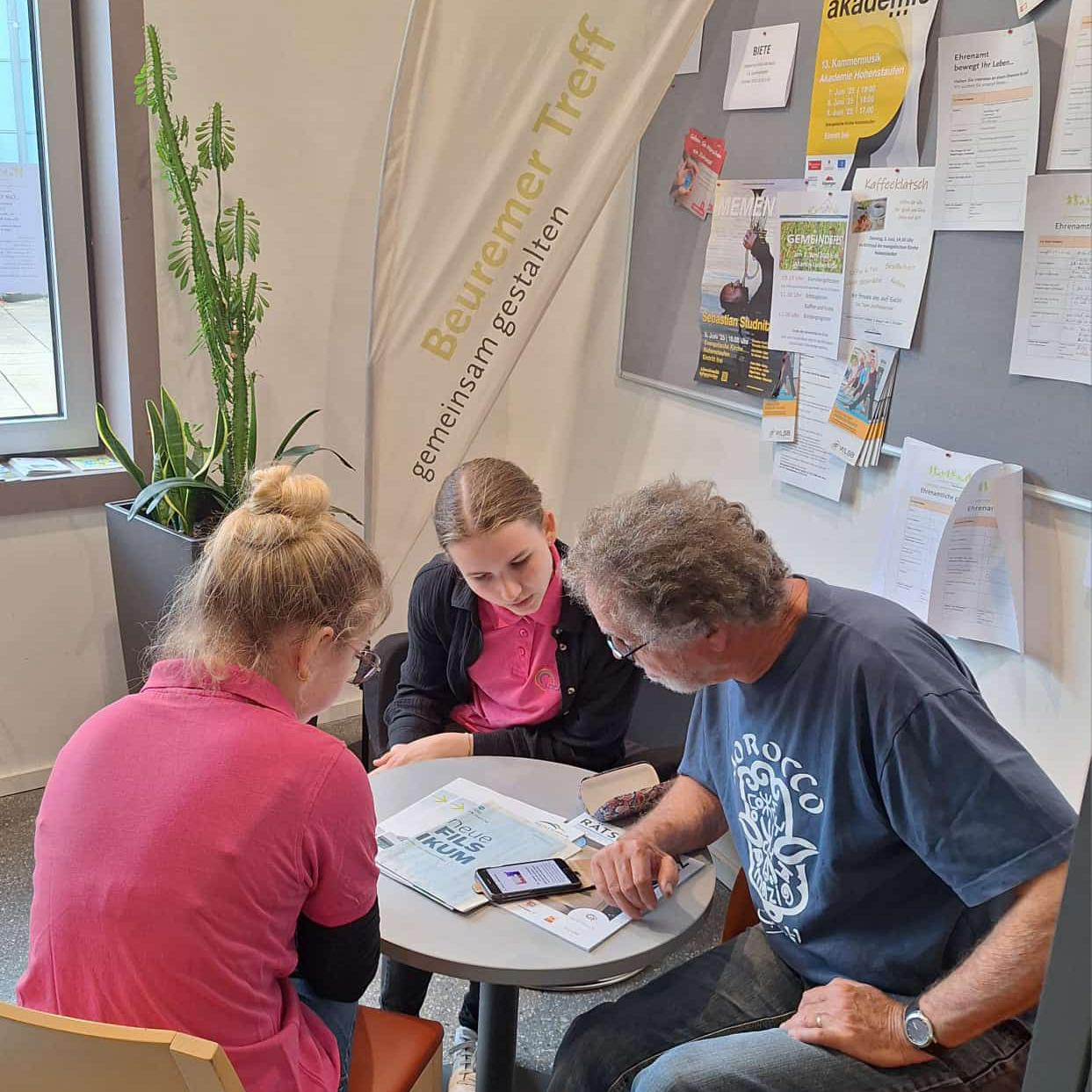 Zwei junge Frauen und ein älterer Mann sitzen um einen kleinen Tisch und schauen auf ein Smartphone und Papiere. Dahinter hängt eine Pinnwand mit Flyern und einem vertikalen Banner mit der Aufschrift „Beuremer Treff Digital-Café gemeinsam gestalten“.