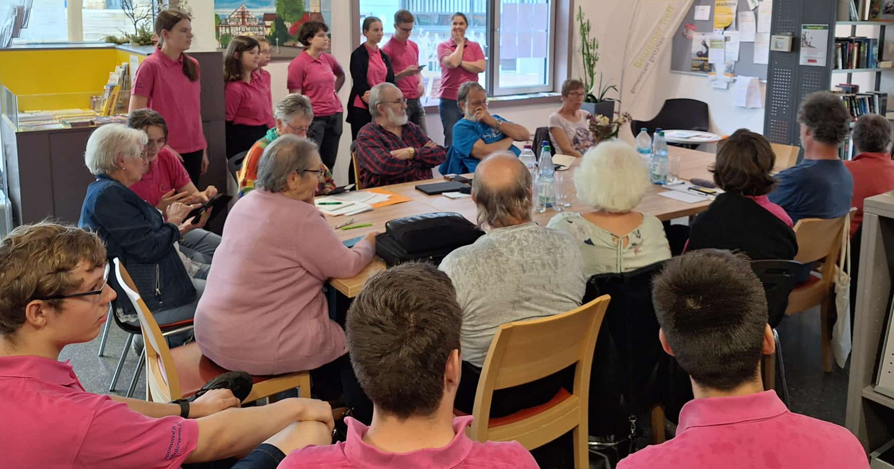Eine Gruppe von Menschen, einige in passenden rosa Hemden, versammelt sich im hell erleuchteten Beuremer Treff zu einer Veranstaltung des Digital-Cafés um Tische und diskutiert. Im Hintergrund sind Bücherregale, Pinnwände und große Fenster zu sehen.