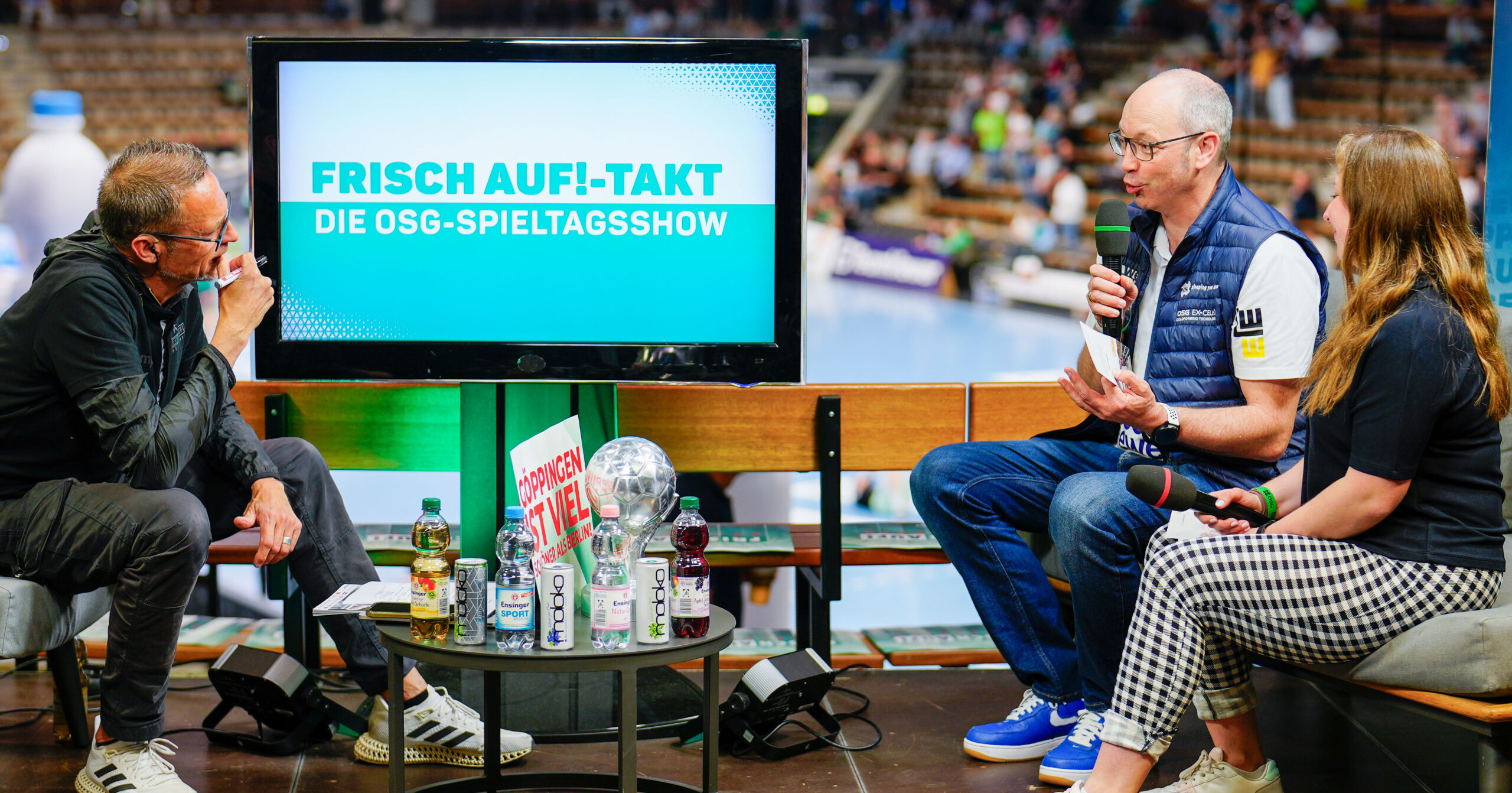 Drei Personen sitzen mit Getränken und Mikrofonen um einen Tisch und diskutieren lebhaft neben einem Bildschirm mit der Aufschrift „Frisch Auf!-Takt Die OSG-Spieltagsshow“ in einer Sportarena – wie ein automatisch gespeicherter Entwurf der Show.