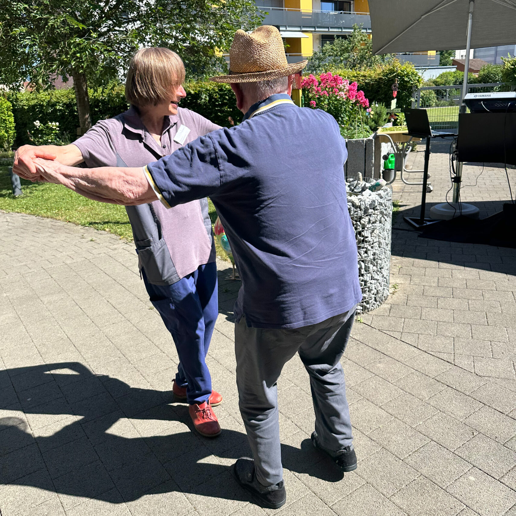Zwei ältere Menschen tanzen an einem sonnigen Tag auf dem Sommerfest in St. Josef, umgeben von Grün und Blumen. Der eine trägt einen Strohhut, der andere rote Schuhe; beide lächeln warm, während sie sich an den Händen halten, mit einem festlichen Gebäude im Hintergrund.