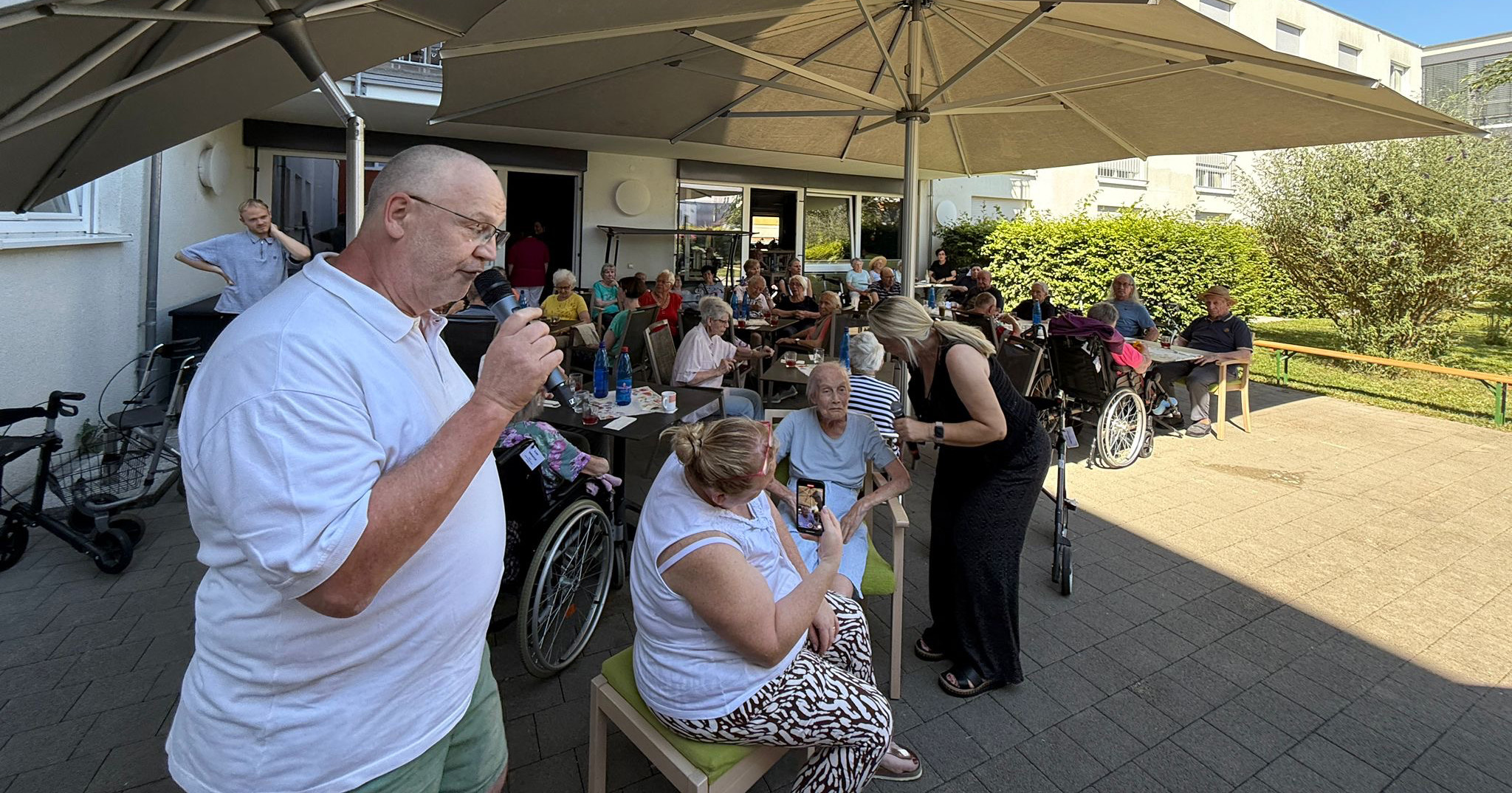 Beim Sommerfest in St. Josef versammeln sich ältere Menschen und ihre Betreuer unter großen Sonnenschirmen, um sich zu unterhalten und an einem sonnigen Tag Eis zu essen. Einige sitzen in Rollstühlen, andere stehen oder sitzen an Tischen und genießen die fröhliche Veranstaltung.