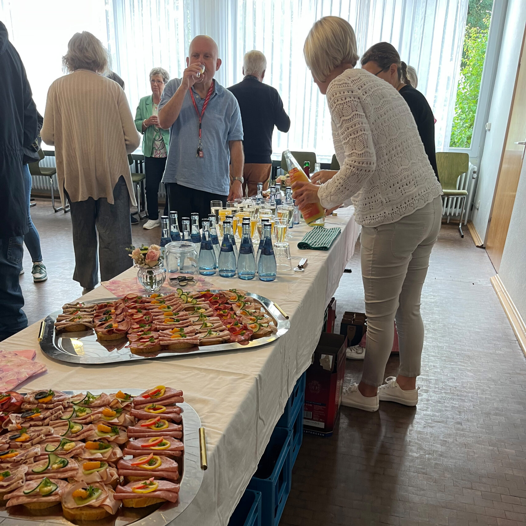 Eine Gruppe versammelt sich um einen langen Tisch in Ellwangen und genießt Tabletts mit belegten Brötchen und Gläsern, während eine Person Orangensaft einschenkt. Während des Christophorustags fällt das Sonnenlicht durch große Fenster.