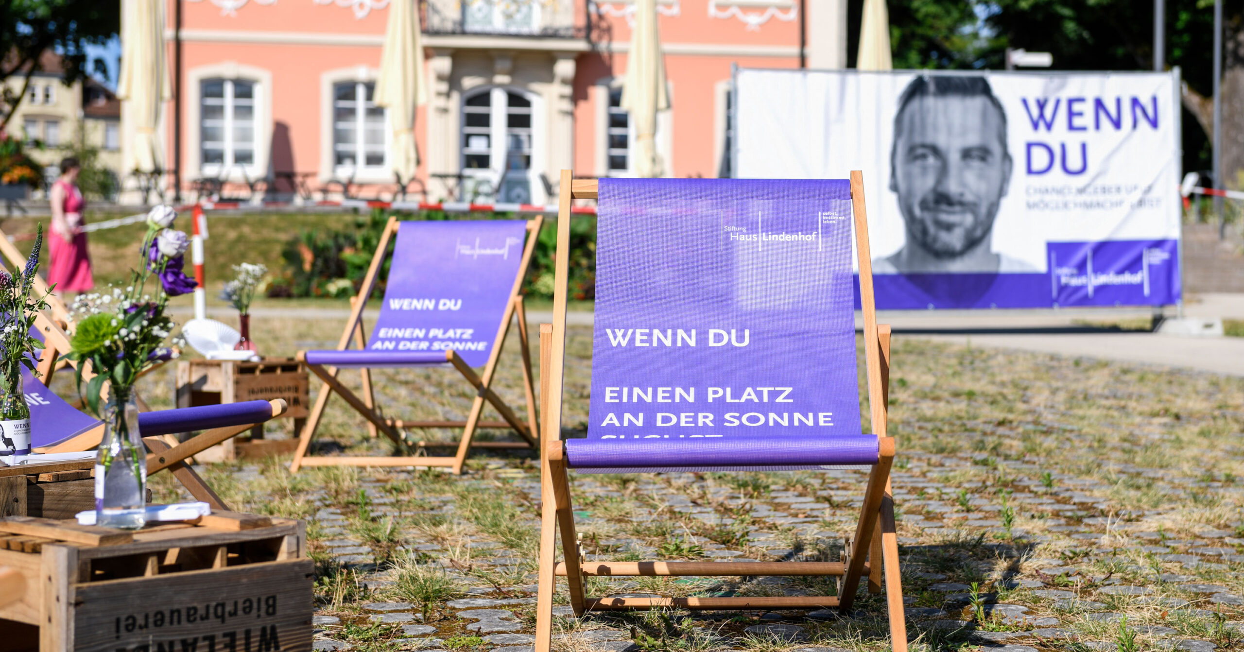 Mehrere lilafarbene Liegestühle mit weißer deutscher Aufschrift stehen im Freien auf Kopfsteinpflaster und erinnern an ein sommerliches und entspanntes Mitarbeiterfest. Im Hintergrund ist ein großes Banner mit einem Männergesicht und einem historischen Gebäude zu sehen.