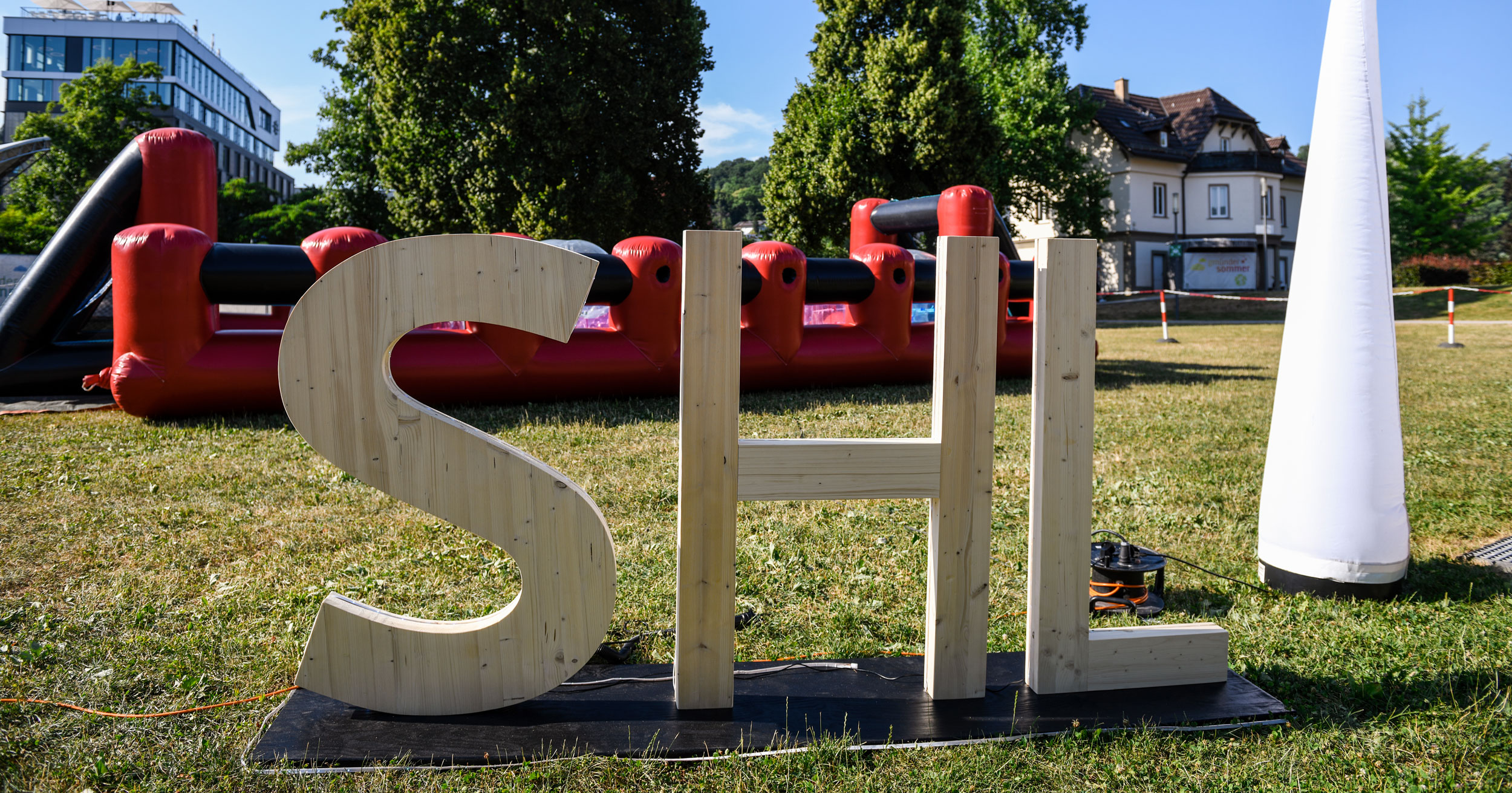 Große Holzbuchstaben mit dem Schriftzug SHL stehen während eines Mitarbeiterfestes auf einer Wiese im Freien, mit aufblasbaren roten Strukturen und Bäumen im Hintergrund unter einem klaren blauen Himmel.