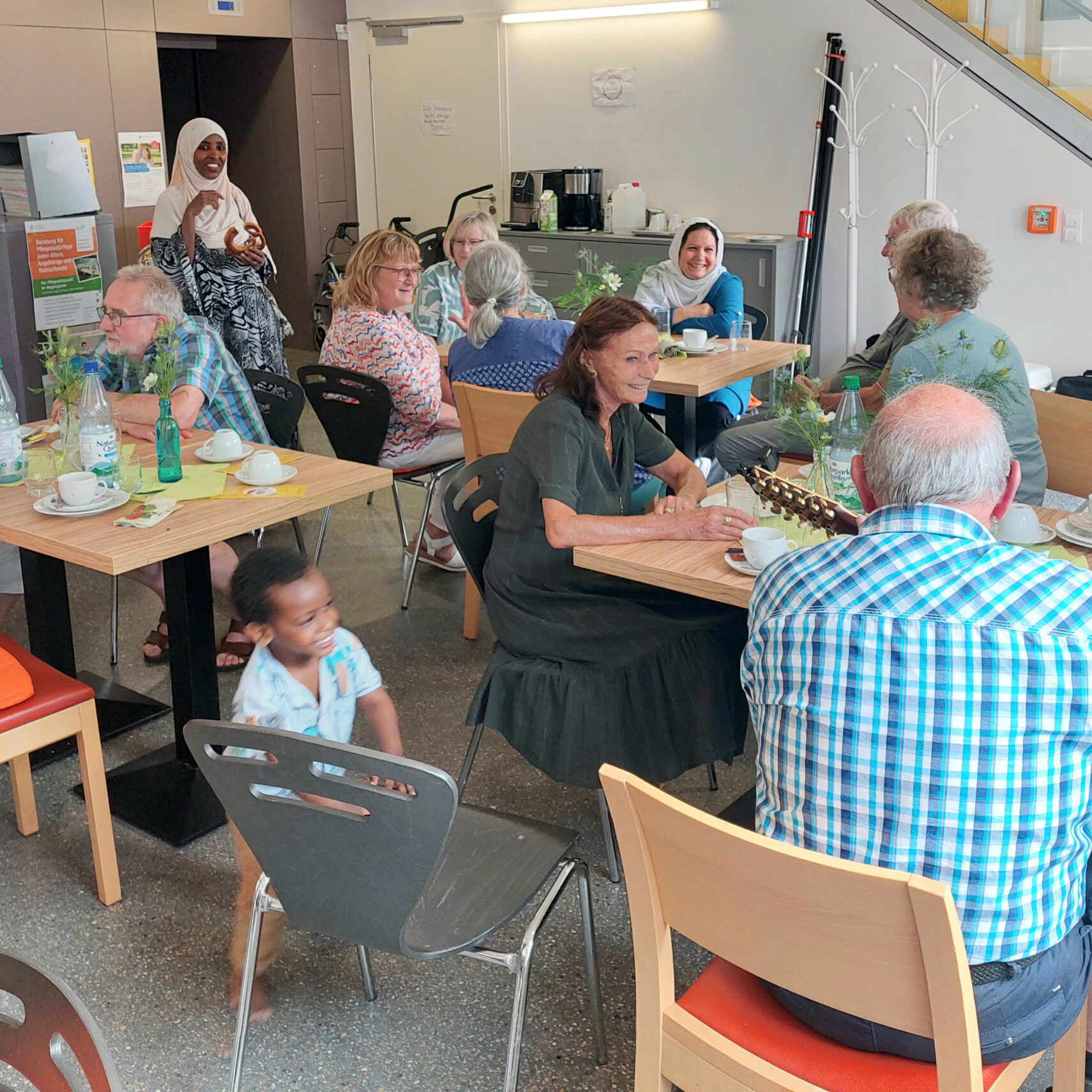 Eine Gruppe von Menschen unterschiedlichen Alters und unterschiedlicher Herkunft sitzt in einem hellen, modernen Café um Tische herum, unterhält sich und lächelt im Café Kunterbunt. Ein kleines Kind geht an den Tischen vorbei, und eine Frau im Hijab steht im Hintergrund und winkt.