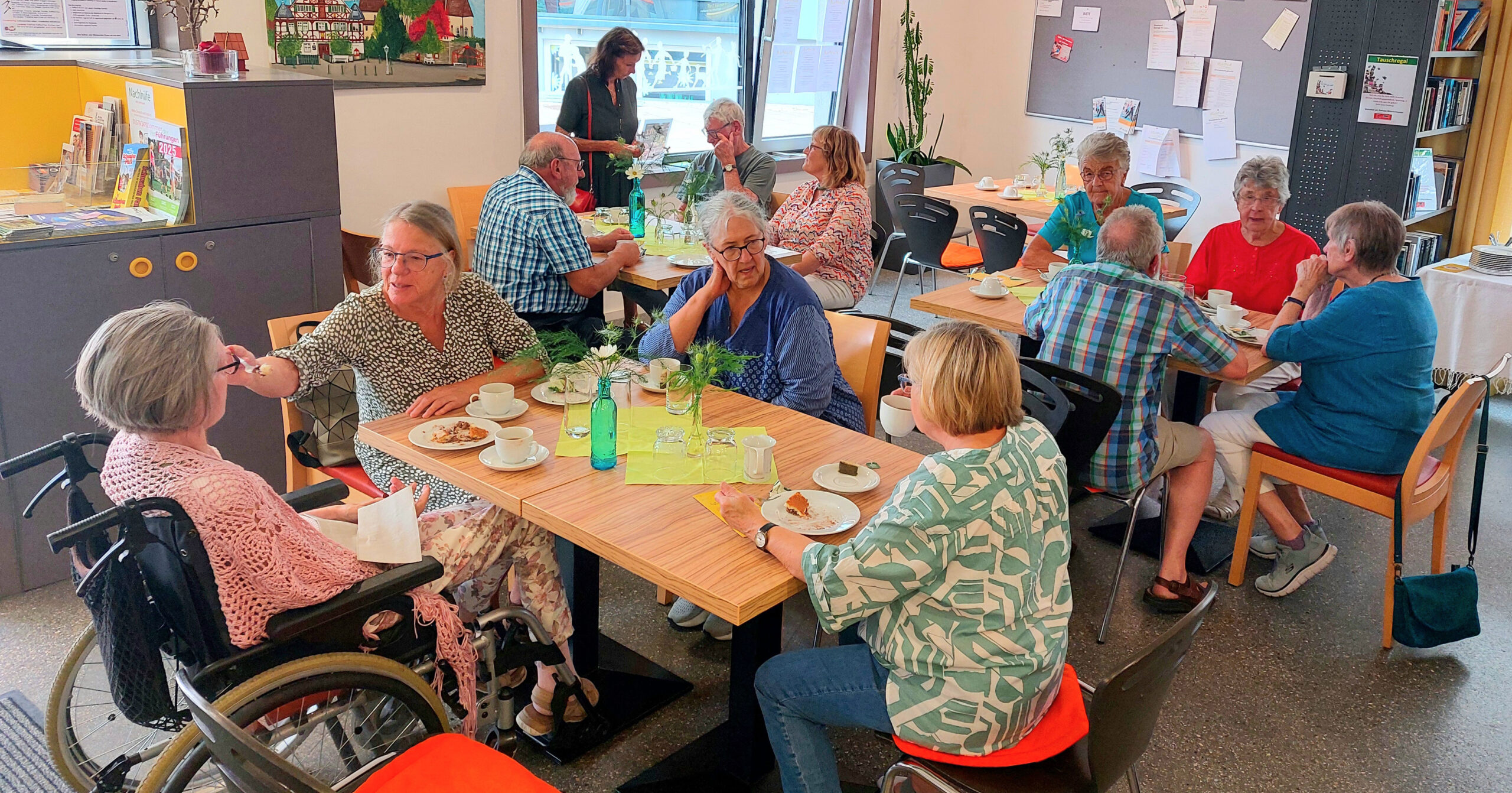 Eine Gruppe älterer Menschen sitzt an Tischen im hellen, ungezwungenen Café Kunterbunt und genießt das Essen und die Gespräche. Eine Frau sitzt in einem Rollstuhl. Die freundliche Atmosphäre ist lebendig, die Menschen lächeln und unterhalten sich.