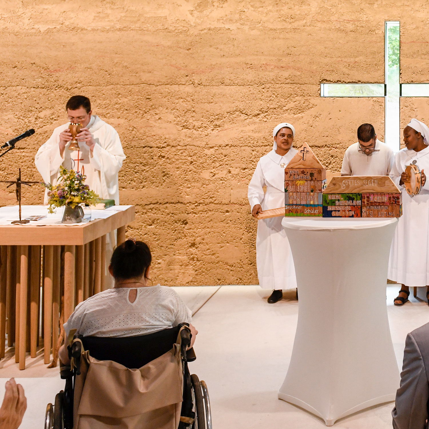 Ein Priester führt eine Einweihungszeremonie am Altar in der Franziskuskapelle durch, während zwei Nonnen und ein Mann einen dekorativen Gegenstand in der Nähe halten; eine Frau im Rollstuhl beobachtet die Zeremonie vom Publikum aus. Im Hintergrund ist ein Kreuz zu sehen.