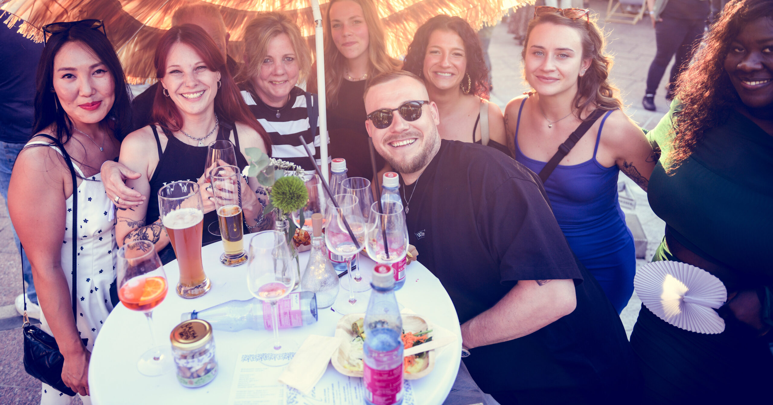 Eine Gruppe lächelnder Freunde, die sich an einem sonnigen Tag auf einem Mitarbeiterfest um einen weißen Tisch im Freien versammelt haben, um Getränke und Essen zu genießen. Einige halten Gläser in der Hand, und ein Regenschirm beschattet einen Teil der Gruppe.