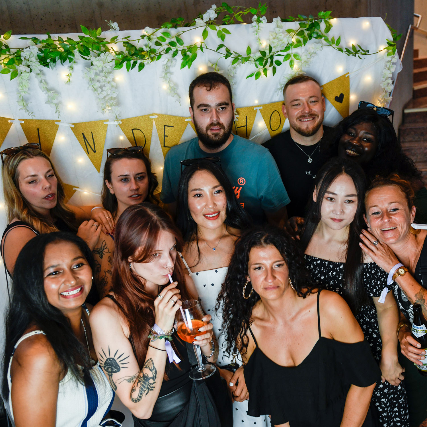 Eine Gruppe lächelnder und posierender Menschen auf einem Mitarbeiterfest, die vor einer dekorierten Kulisse mit Lichtern, Grünzeug und einem Banner stehen. Einige halten Getränke in der Hand, und alle sind lässig und sommerlich gekleidet.