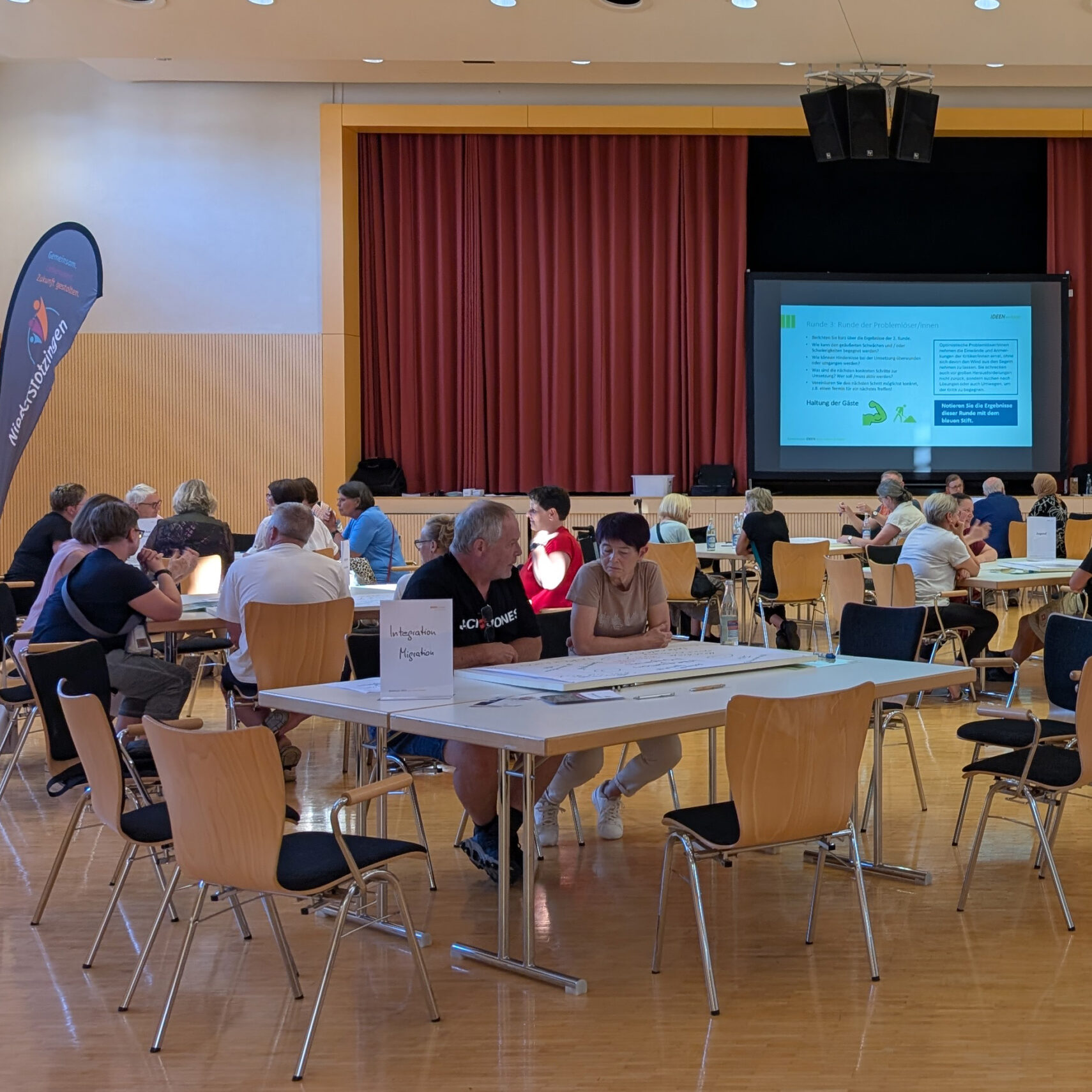 In einem großen Raum mit Holzfußboden sitzen Menschen in Gruppen an Tischen und arbeiten gemeinsam in der Ideenschmiede Niederstotzingen. Vorne zeigt ein Beamer Informationen an, dahinter ein roter Vorhang, auf einigen Tischen sind Schilder zu sehen.