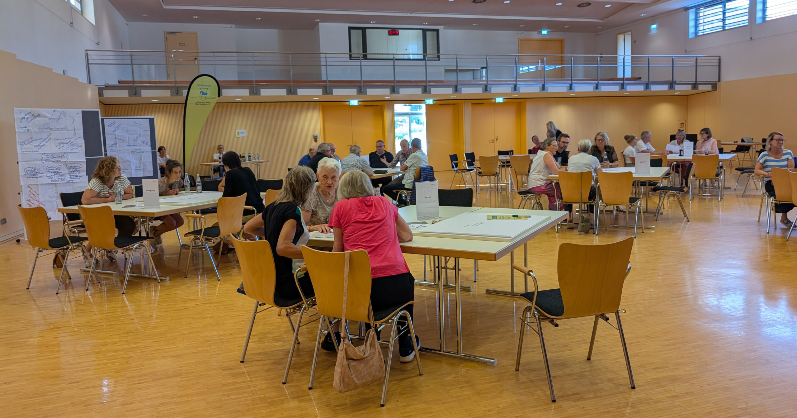 In einem geräumigen, hellen Raum mit Holzfußboden sitzen Menschen in kleinen Gruppen an Tischen, unterhalten sich und arbeiten. Auf der linken Seite sind Wandkarten und Papiere ausgestellt - eine Ideenschmiede-Atmosphäre in Niederstotzingen - und ein paar Leute laufen herum.