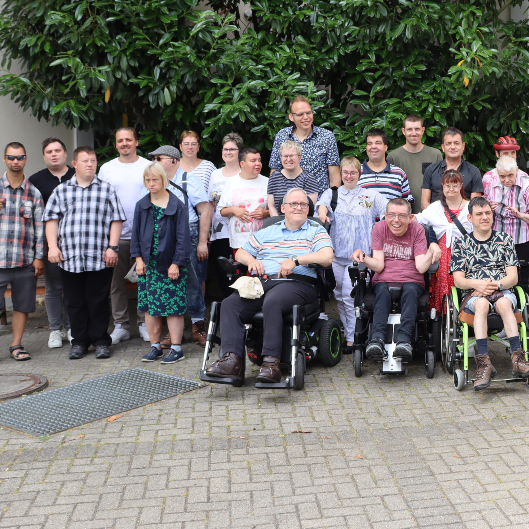 Eine Gruppe Erwachsener, teils stehend, teils in Rollstühlen, posiert während einer Jubiläumsfeier in Ellwangen vor einem belaubten Baum und lächelt in die Kamera auf einer gepflasterten Fläche in der Nähe eines Gebäudes.