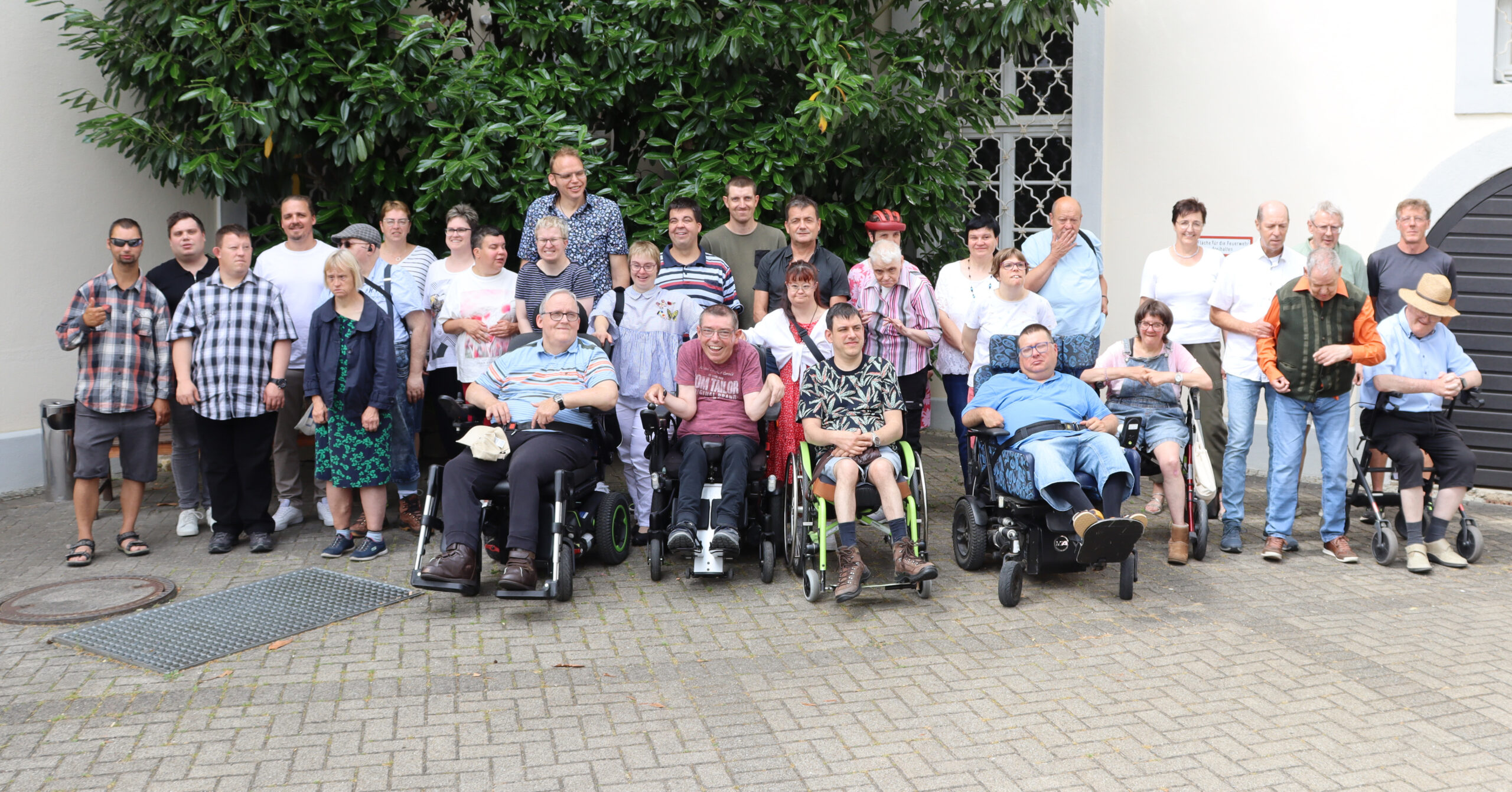 Eine große Gruppe Erwachsener, einige im Rollstuhl, andere im Stehen, versammelt sich zu einer Jubiläumsfeier in Ellwangen und posiert im Freien vor einem grünen Baum und einem weißen Gebäude, alle lächeln in die Kamera.
