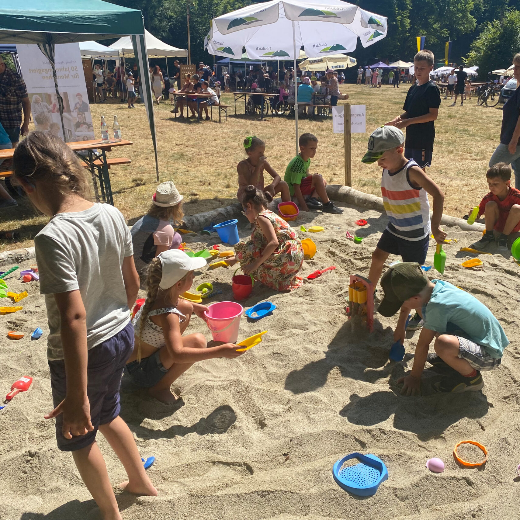 Kinder spielen mit Sandspielzeug in einem großen Sandkasten im Freien bei einer sonnigen Veranstaltung, umgeben von Zelten und Menschen. Einige Kinder graben, während andere mit Eimern und Schaufeln hantieren. Ihre Kreativität formt einen automatisch gespeicherten Entwurf von Spaß.